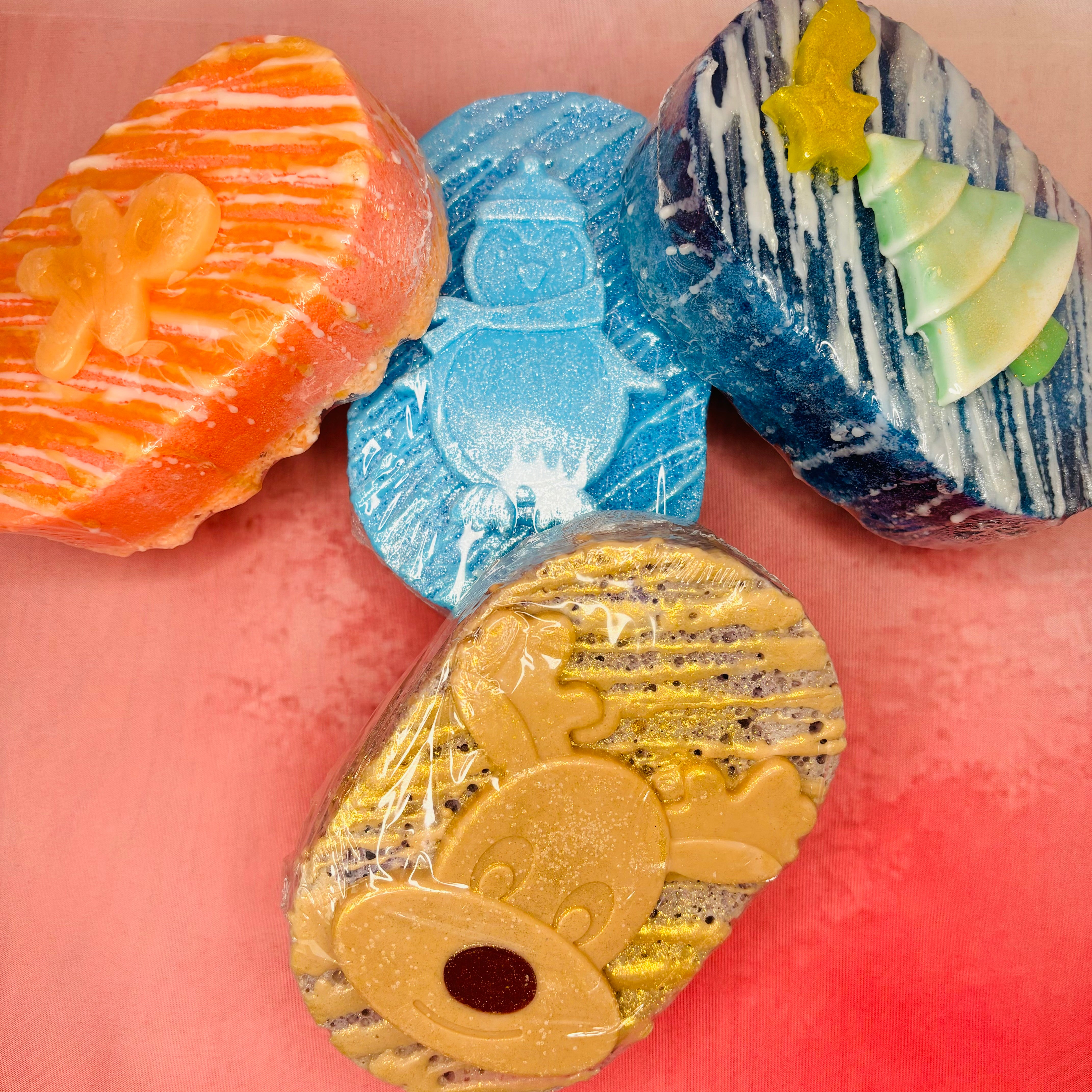 Four vibrant soap sponges from The Soap Gal x, including a Gingerbread Man Soap, with festive designs featuring gingerbread, penguin, tree, and reindeer shapes—offer a luxurious lather. These are creatively displayed against a pink background.