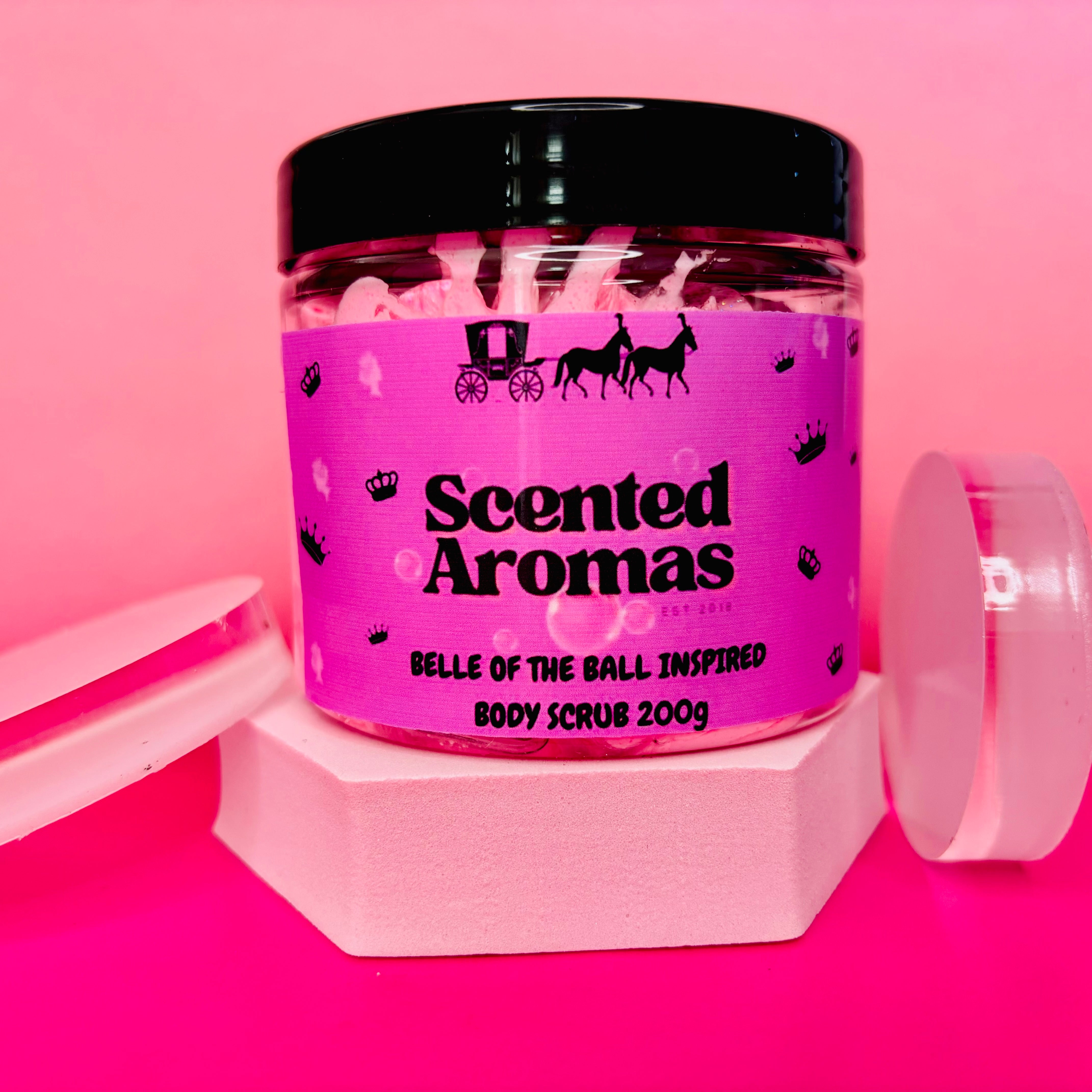 A jar of The Soap Gal x "Belle Of The Ball Sugar Body Scrub 200g," featuring a pink label and black lid, is elegantly placed on geometric pink shapes against a pink background. It offers a delightful fragrance while promising to exfoliate your skin with finesse.