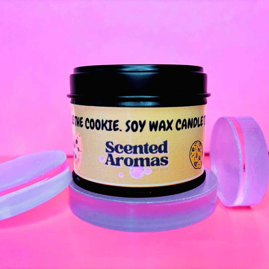 A black jar labeled "The Soap Gal x" containing the Baked Chocolate Cookies Candle Tin - 100ml is placed among translucent circular objects on a pink background. This soy wax candle offers to transform your space into a deliciously cozy haven.