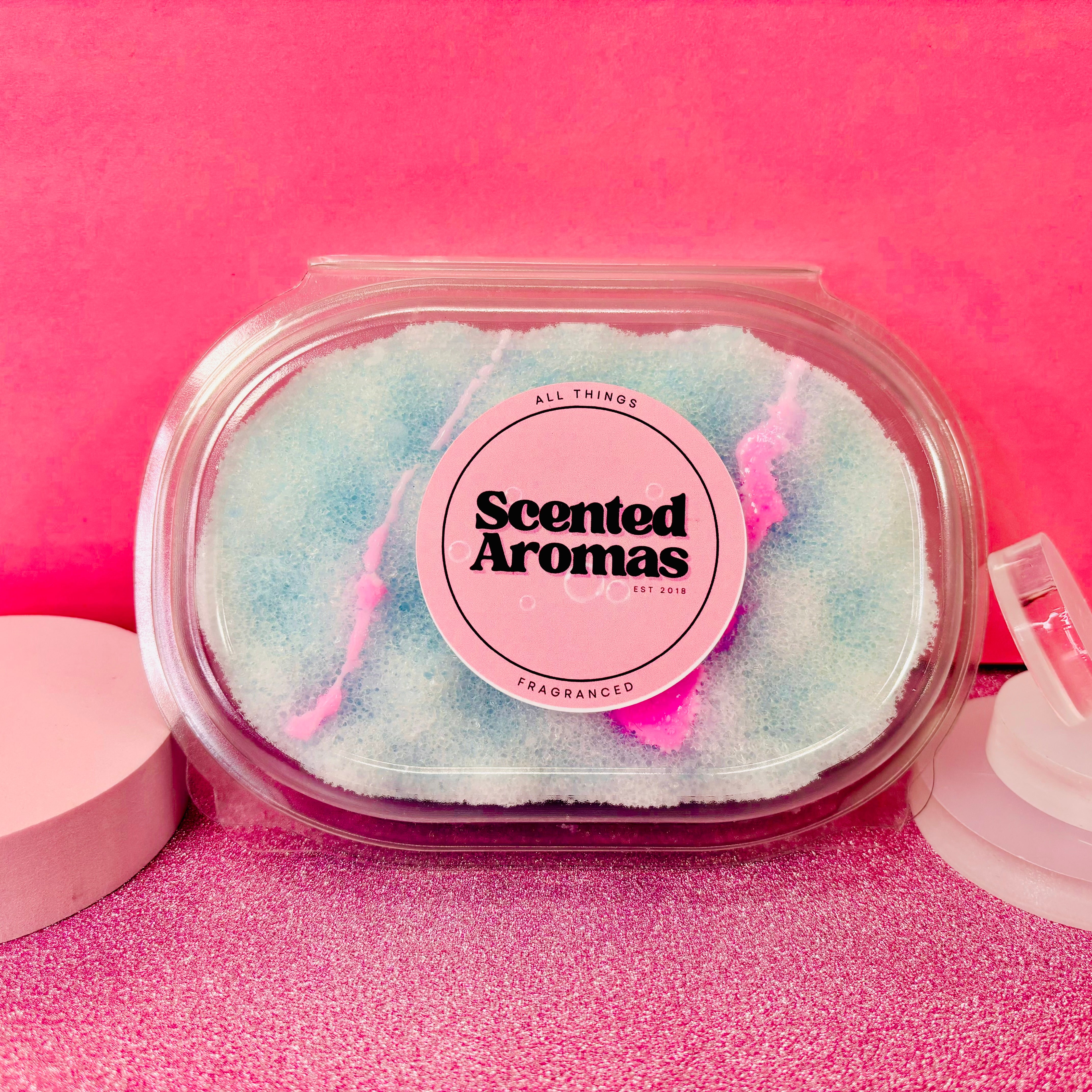 A container of scented crystals labeled "Scented Aromas" rests gracefully against a pink background, with its blue and pink hues reminiscent of a serene skincare routine. Perfectly complementing your Baby Powder Exfoliating Soap Sponge by The Soap Gal x, these crystals can elevate your soapy sponge experience to new heights.