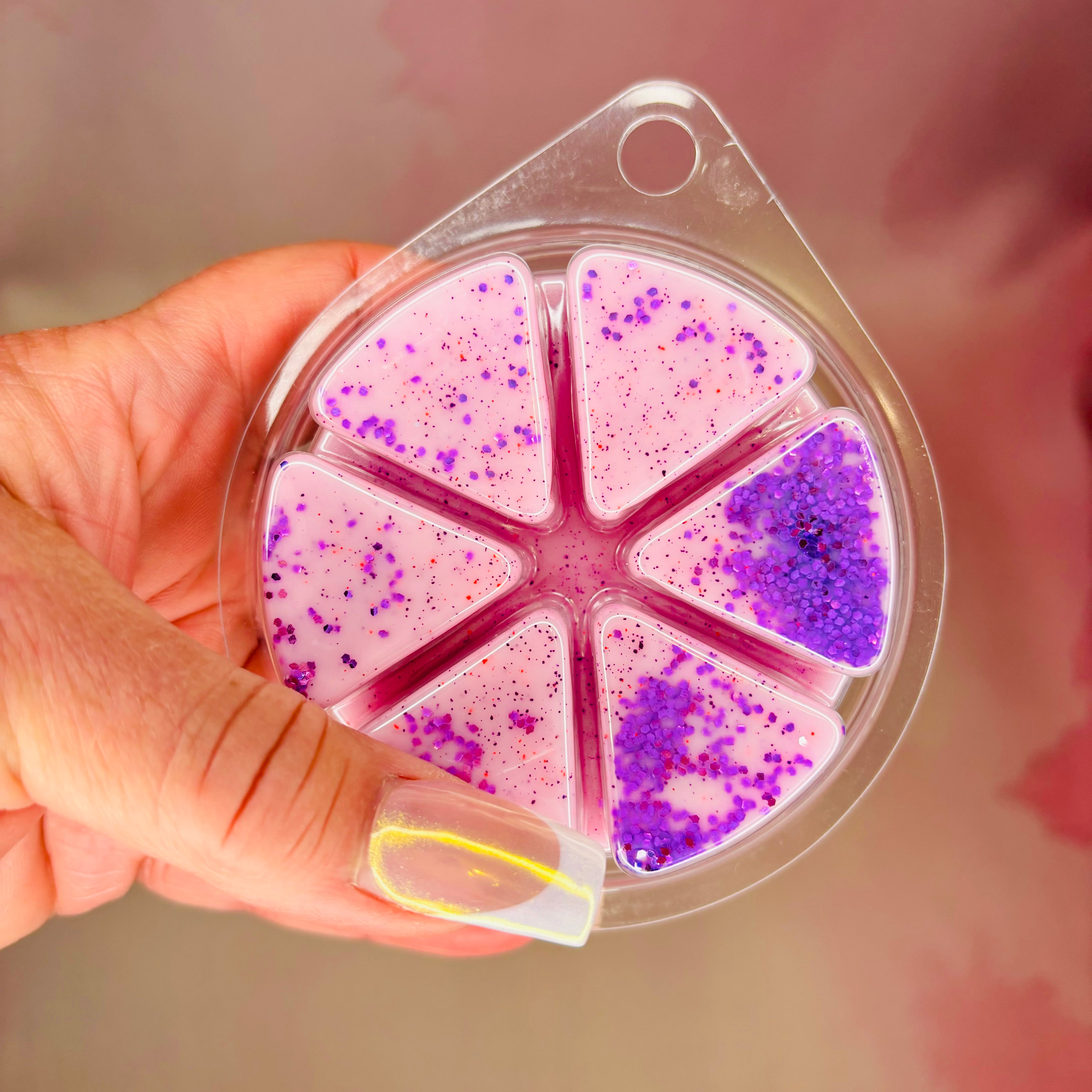 A hand holds a circular container with six compartments filled with the Alien Encounter Wax Melt by The Soap Gals, its biodegradable pink and purple glitter shimmering like cosmic stardust.