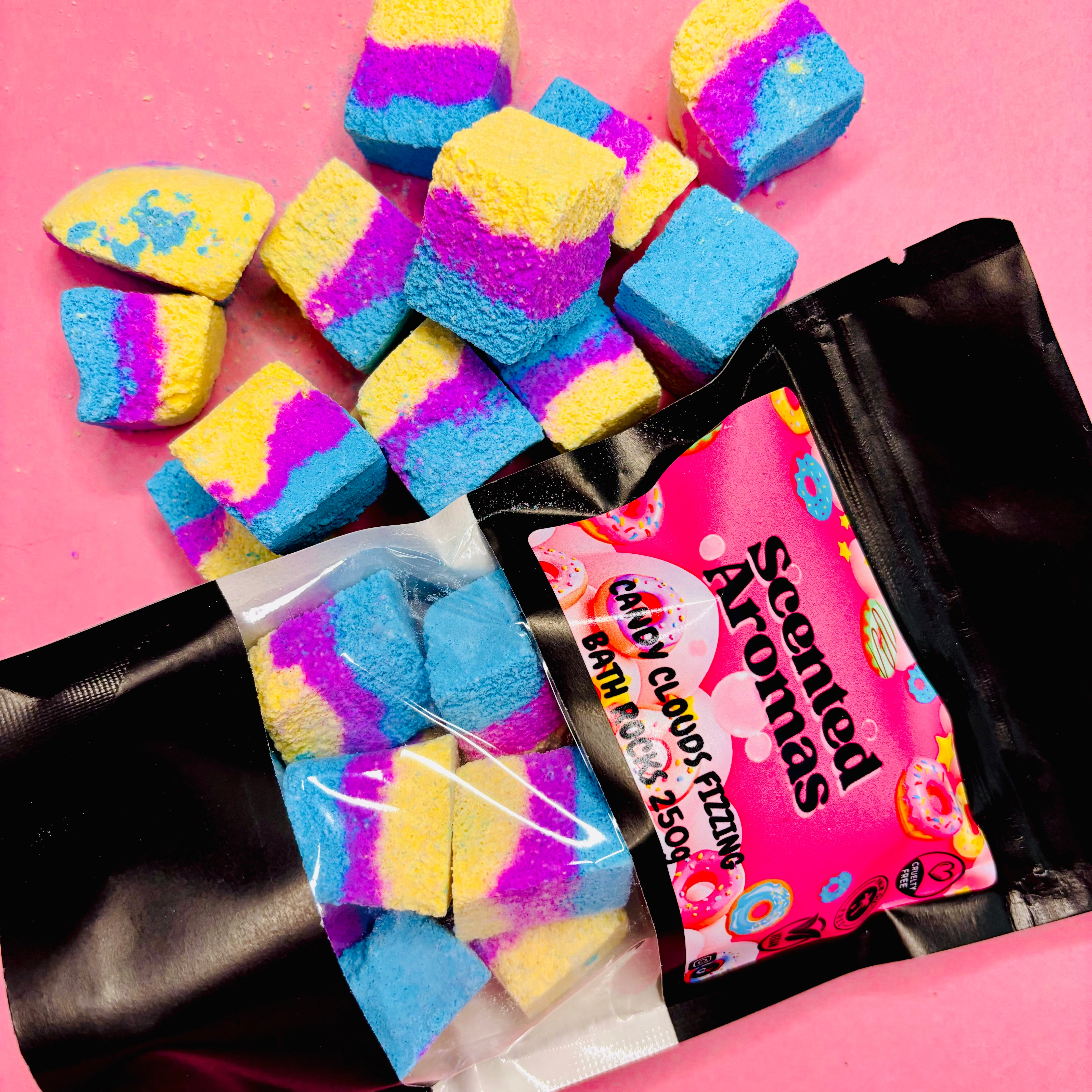 Bath Bomb Rocks in blue, yellow, and pink tumble out of an open black package labeled "The Soap Gal x" on a pink background. This colorful display evokes the whimsical charm of Cotton Candy and Rainbow Sprinkles.