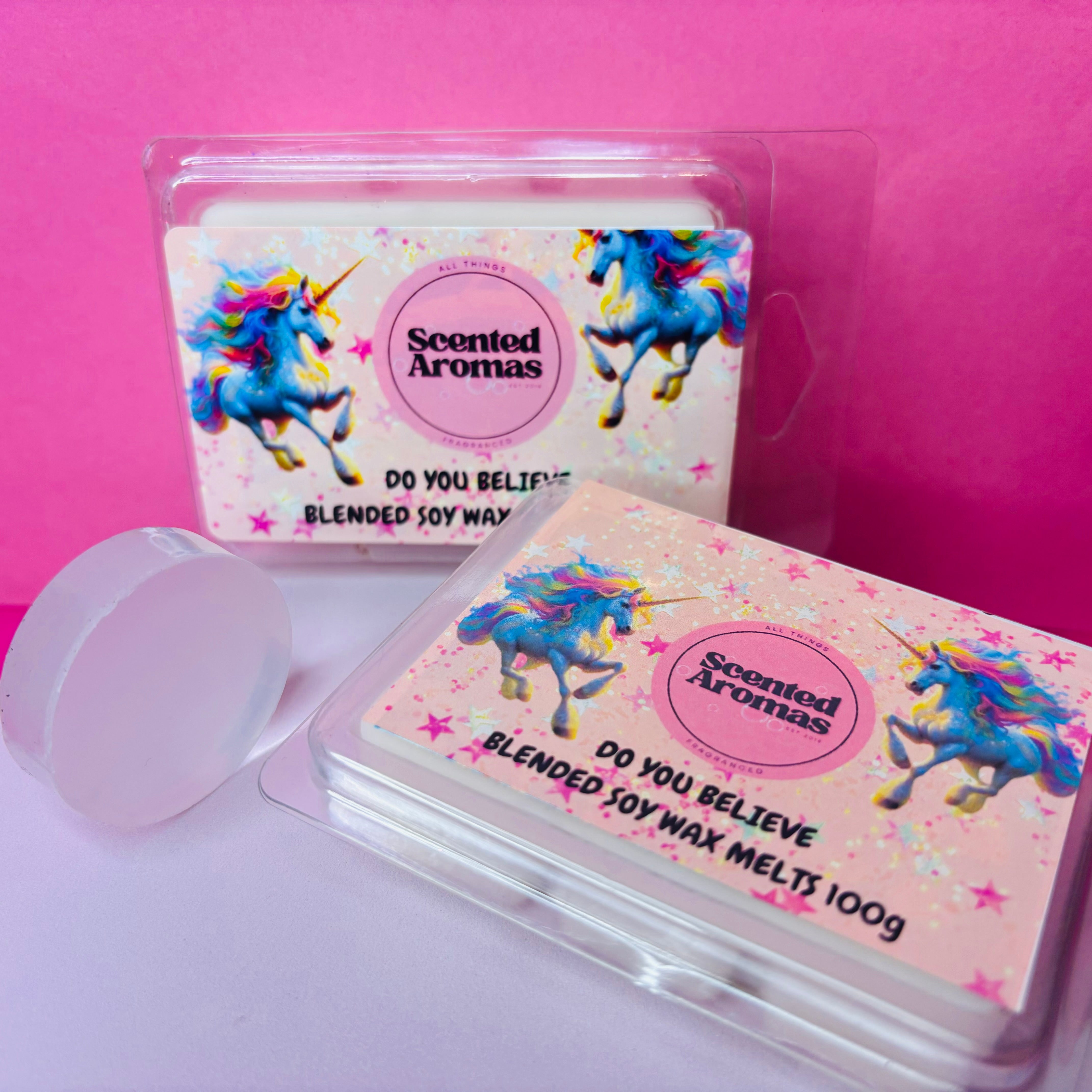 Two packs of "Do You Believe In Magic Wax Melts" by The Soap Gal x, featuring unicorn-themed labels and emanating a captivating vanilla musk fragrance. One wax melt is out of its packaging, set against a pink and white backdrop.