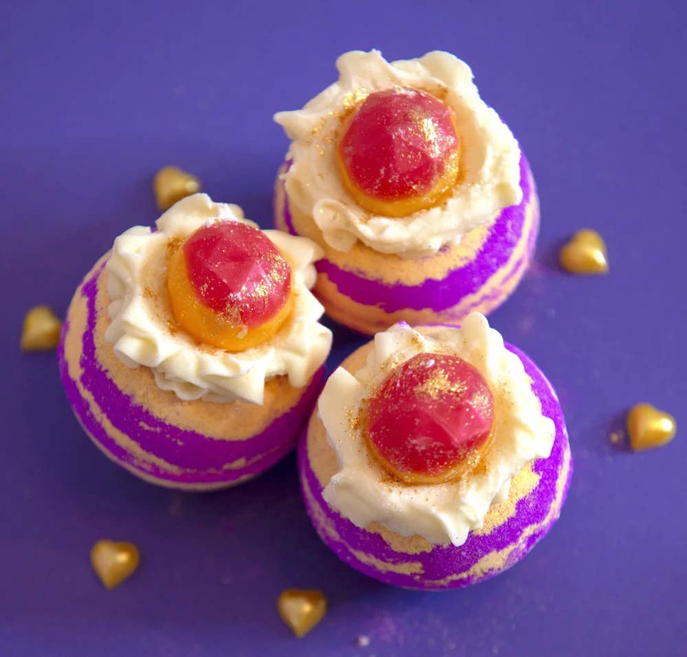 Three Rhubarb & Custard Cocoa Shea Butter Bath Bombs from Scented Aromas, adorned with creamy white frosting and red jelly-like tops, rest elegantly on a purple surface. Each one is sprinkled with Bio Glitter and small gold heart decorations, adding an extra touch of indulgence to your bath time.