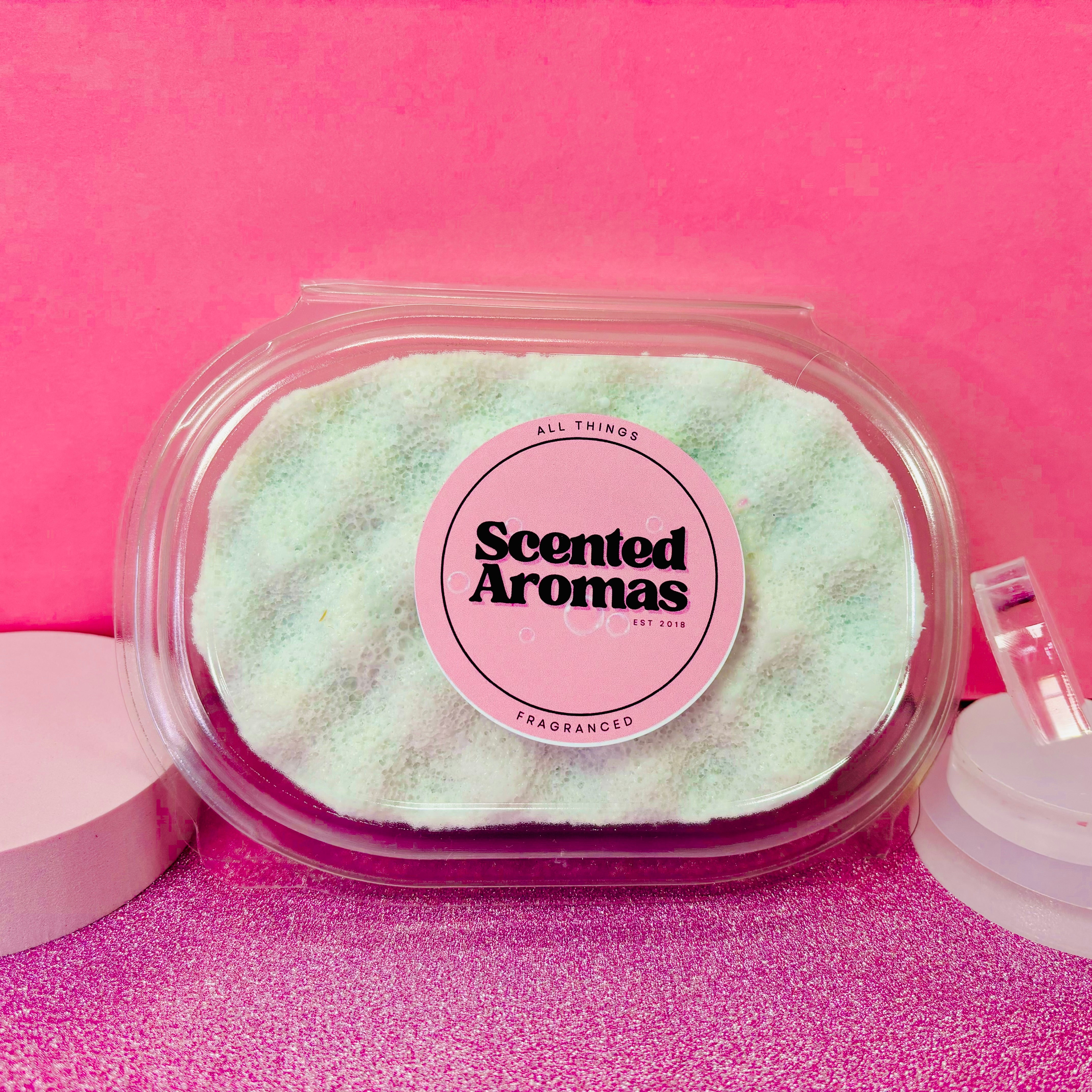 A clear container featuring The Soap Gal x Kreed Exfoliating Soap Sponge, labeled "Scented Aromas," set against a pink background, makes for an ideal enhancement to any men's skincare routine.