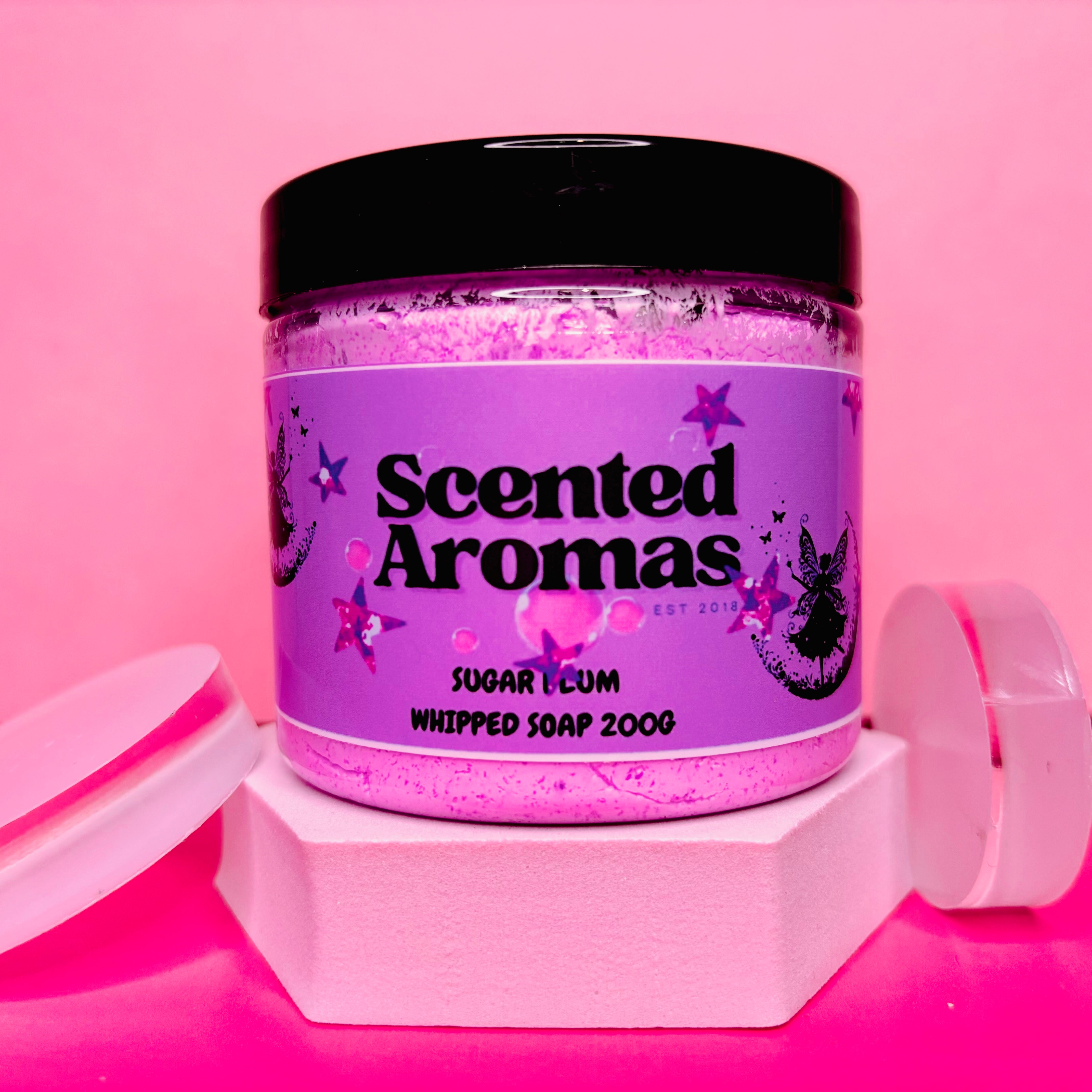 A jar of The Soap Gal x Frosted Sugar Plum Whipped Soap Shave Butter, 200g, infused with a sweet and frosty scent, is elegantly placed on a pink hexagonal stand. The accompanying pink lid enhances the vibrant background, offering moisturizing and skin-hydrating benefits with every application.