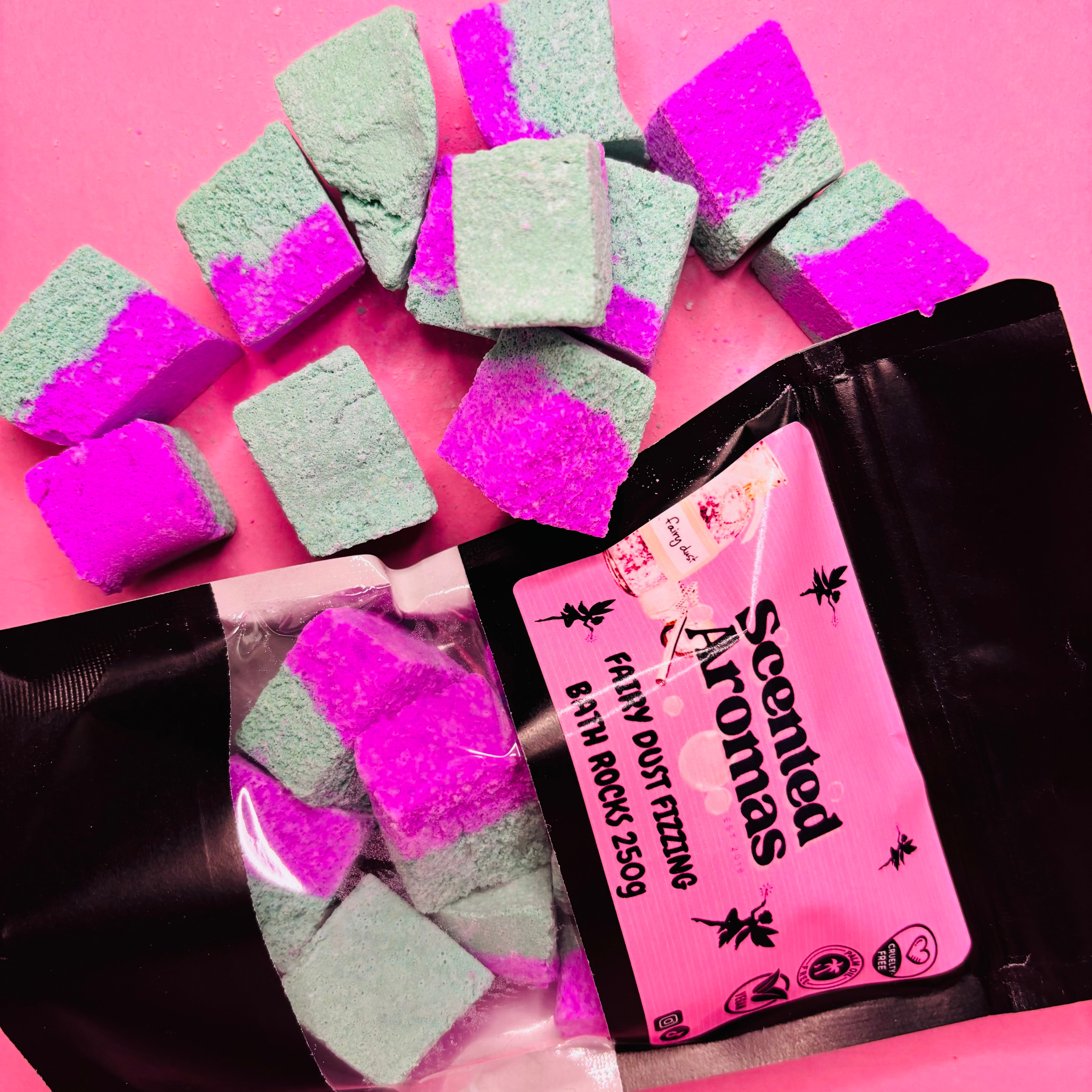 Pink and green bath rocks are artfully spread over a pink surface, accompanied by a labeled bag that reads "The Soap Gal x A Fairies Wish Bath Bomb Rocks 250g." These charming bath rocks provide a luxurious experience reminiscent of Pink Prosecco's sparkling allure.