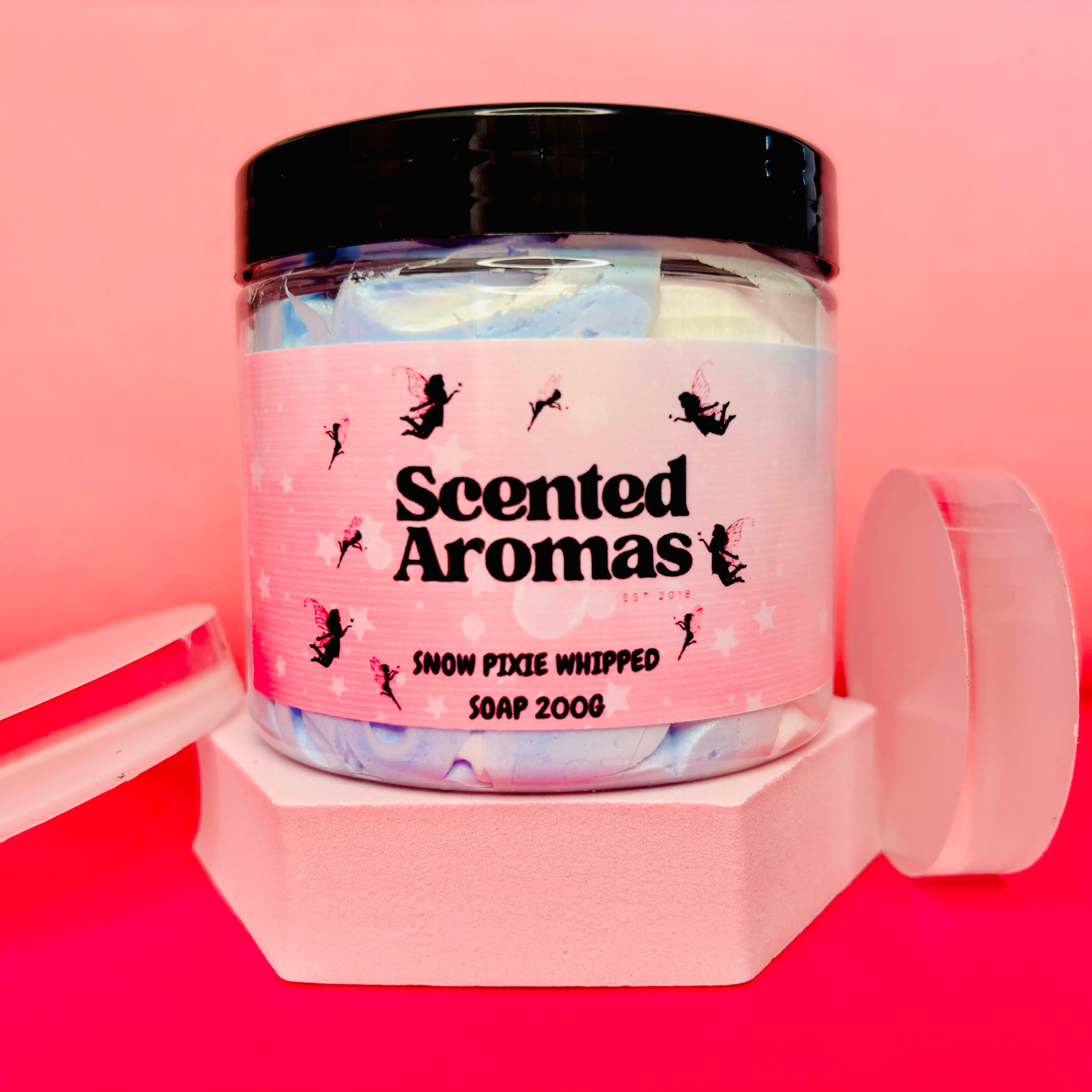 A 200g jar of The Soap Gal x Snow Pixies Whipped Soap Shave Butter features a sweet scent, complemented by a pink label adorned with fairy graphics and set against a pink background.