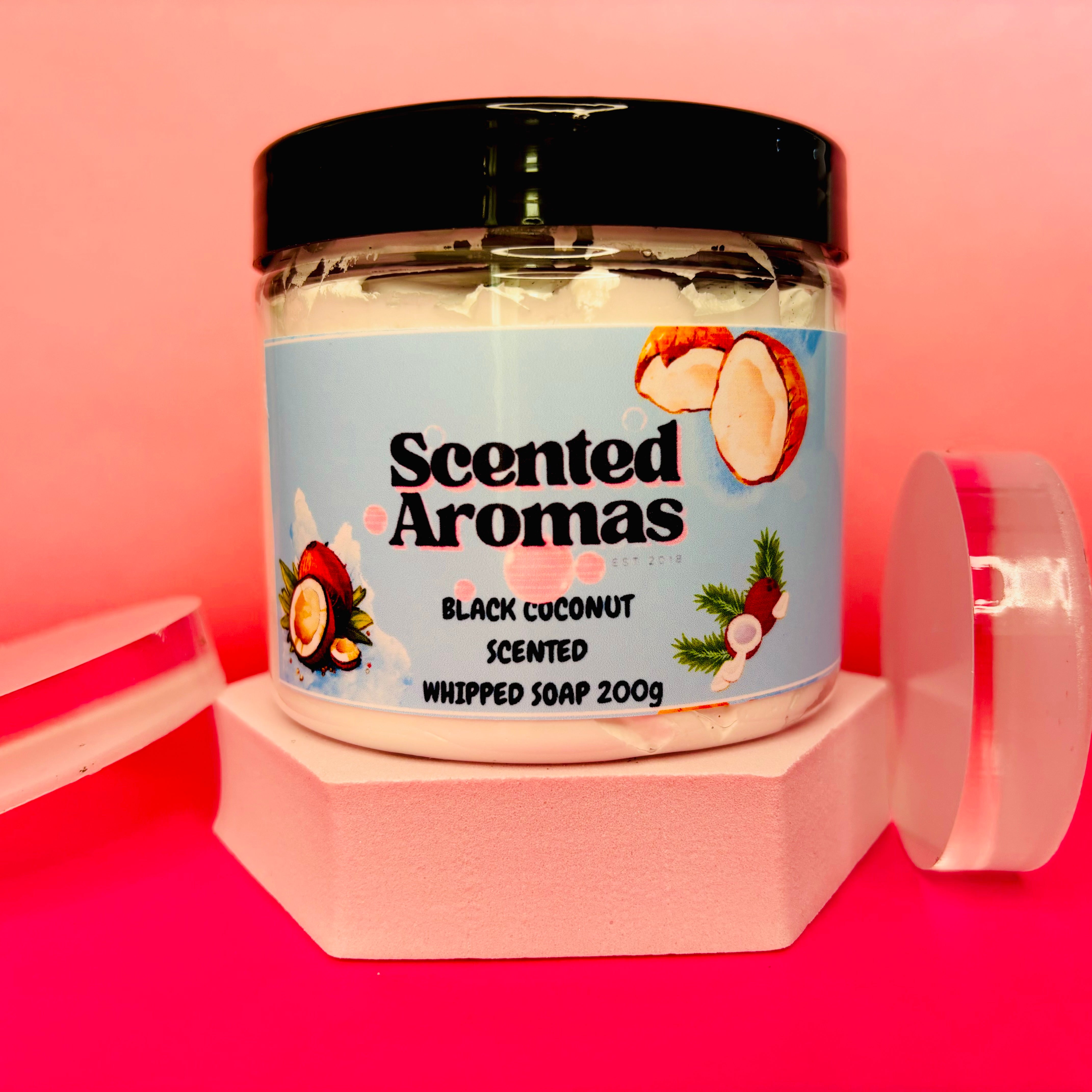 A jar of The Soap Gal x "Black Coconut Whipped Soap Shave Butter," ideal for your skincare routine at 200g, is set against a pink background with soap bars beside it.