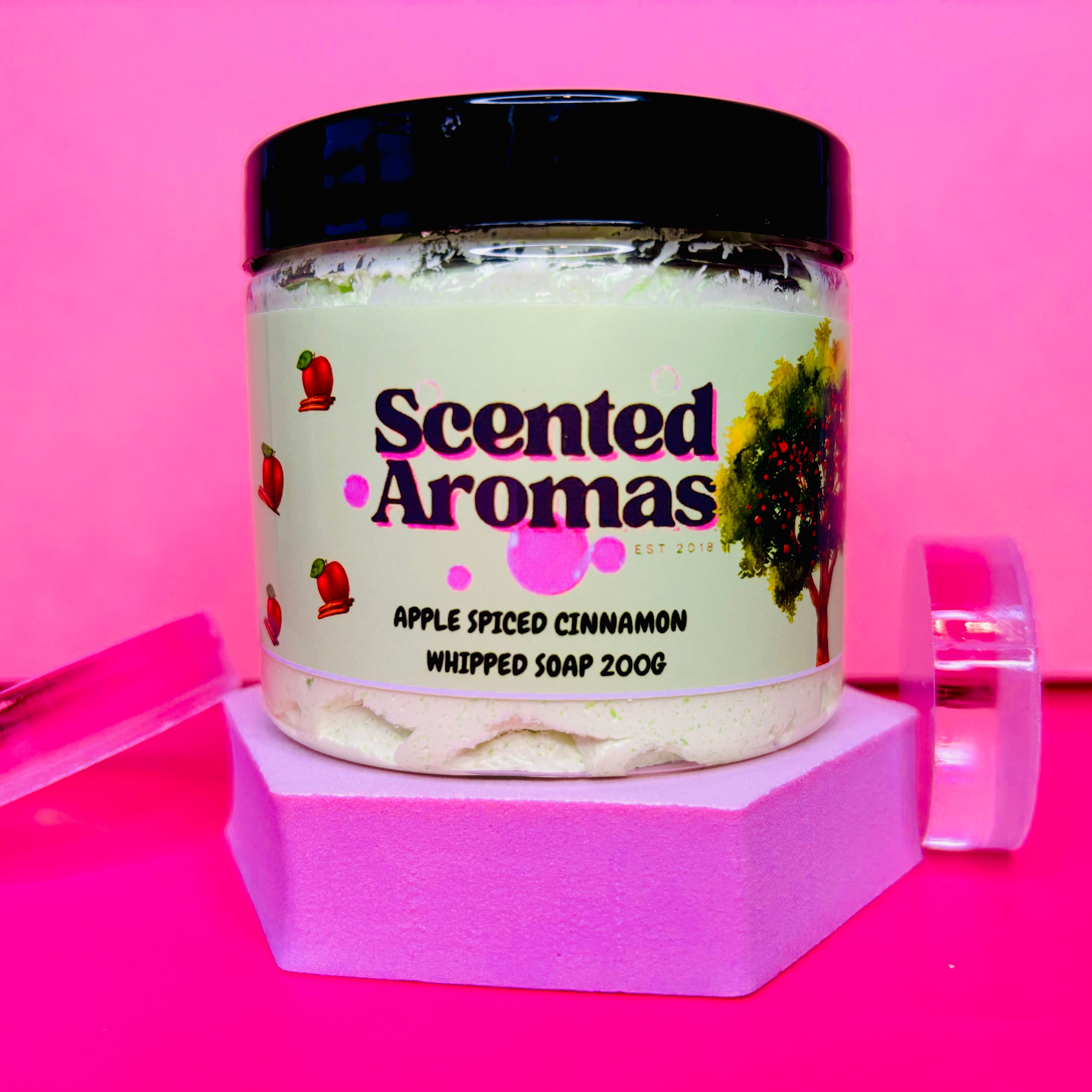 A jar of The Soap Gal x Cinnamon and Apple Whipped Soap Shave Butter, 200g, is elegantly displayed on a pink hexagonal stand against a bright pink background, making it perfect for winter skincare.