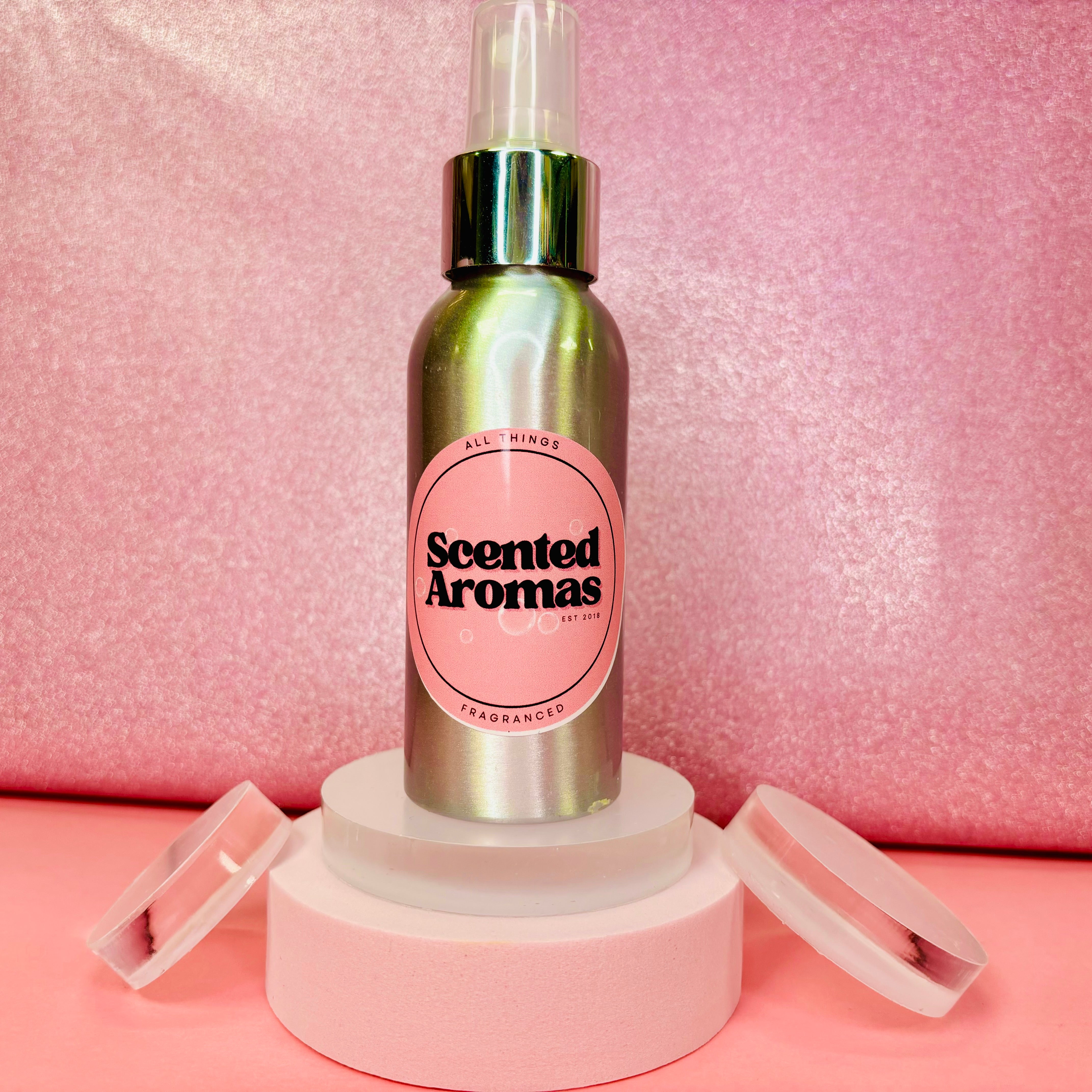 A Baccarat Inspired Body Spray from The Soap Gal x, elegantly displayed on a round pedestal, sits amidst two open lids against a pink textured background, radiating a sophisticated aroma.