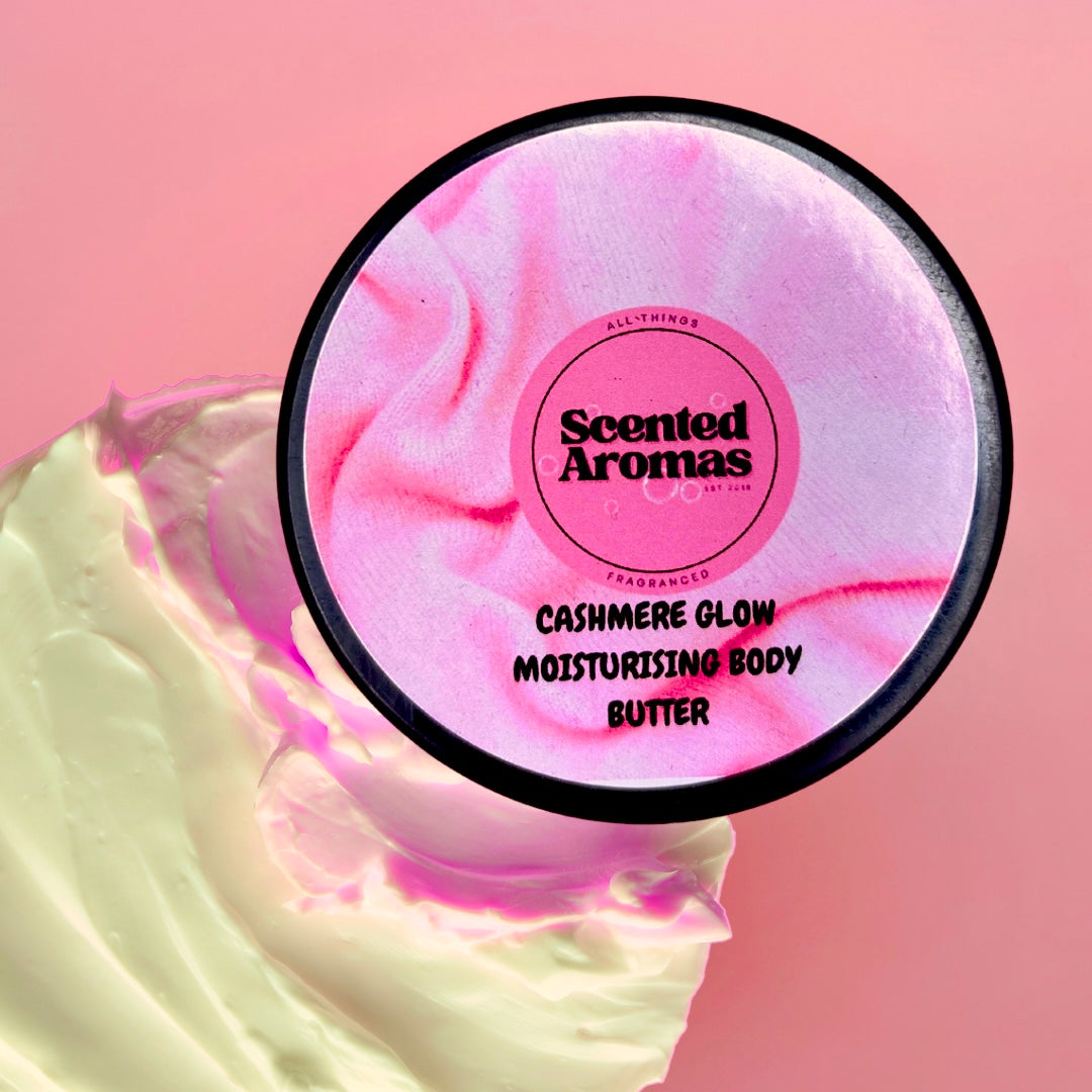 A round container labeled "The Soap Gal x Wrapped In Cashmere Body Moisturising Butter 100ml" rests against a pink background, with its swirled cream texture suggesting luxurious hydration.