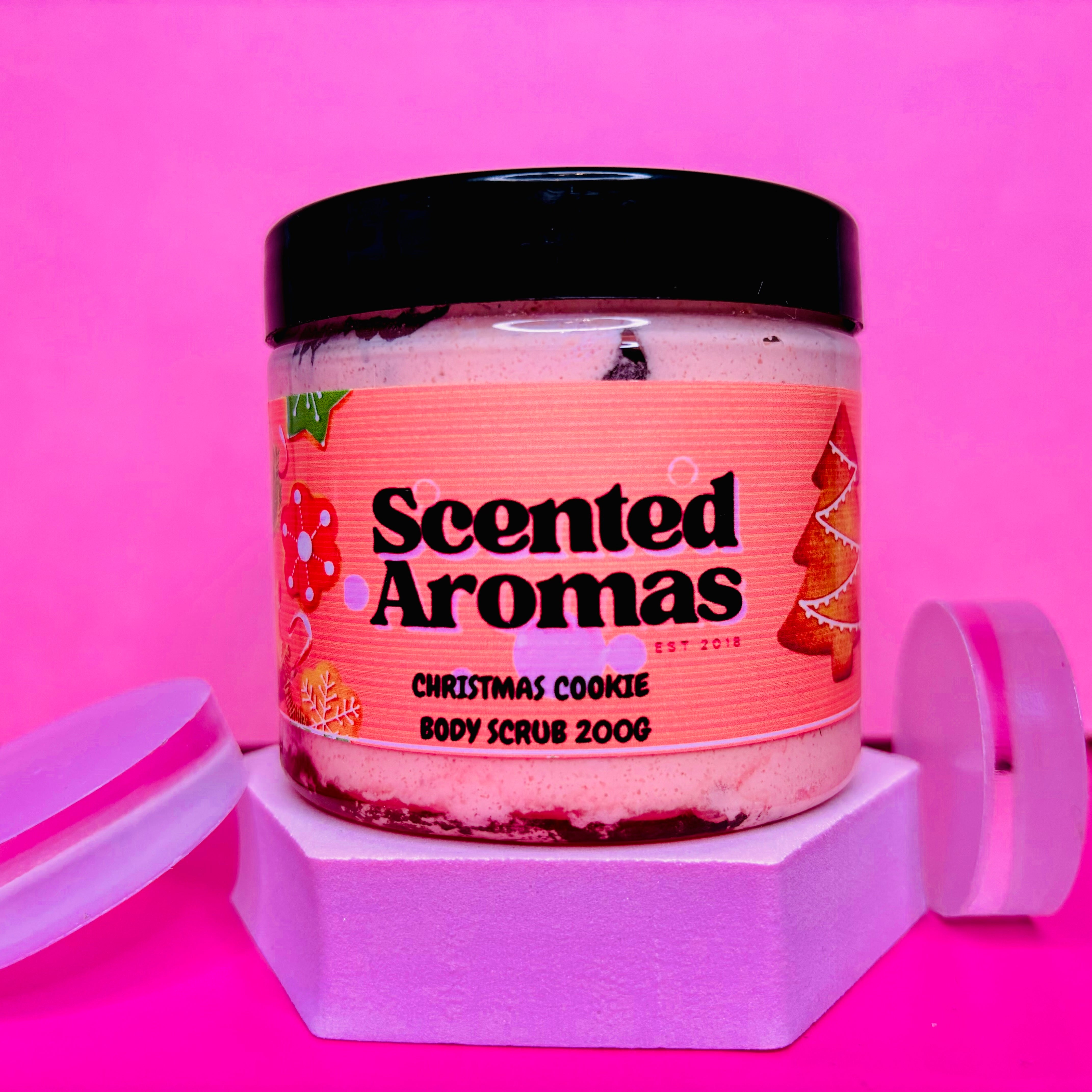 A jar of "The Soap Gal x" Christmas Cookies Sugar Body Scrub, 200g, featuring a pink label, is displayed on a hexagonal stand against a pink background, ideal for achieving that winter-ready glow.