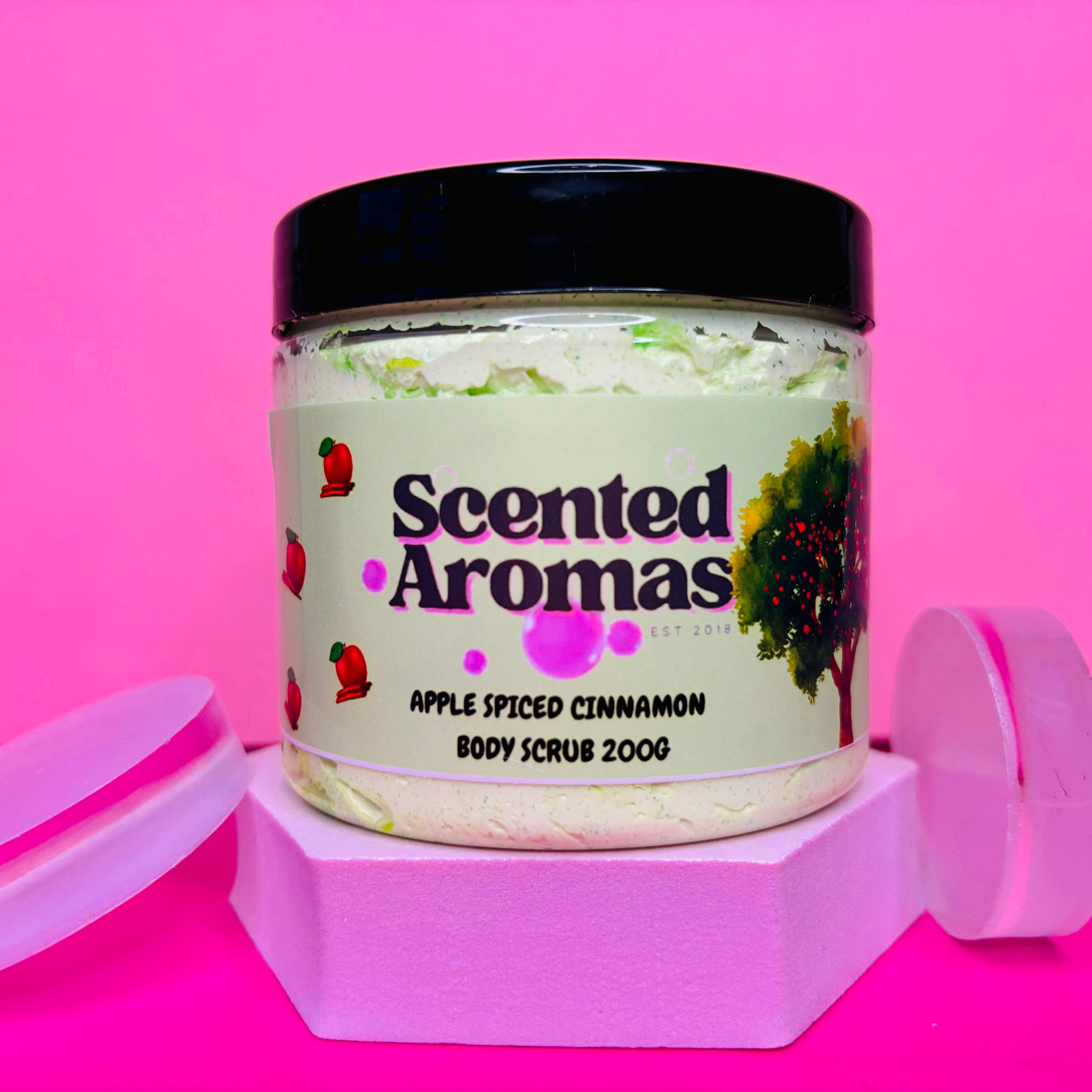 A jar of Cinnamon and Apple Sugar Body Scrub 200g by The Soap Gal x is displayed on a hexagonal stand against a pink background, crafted as an exfoliating sugar scrub that also enhances skin circulation.