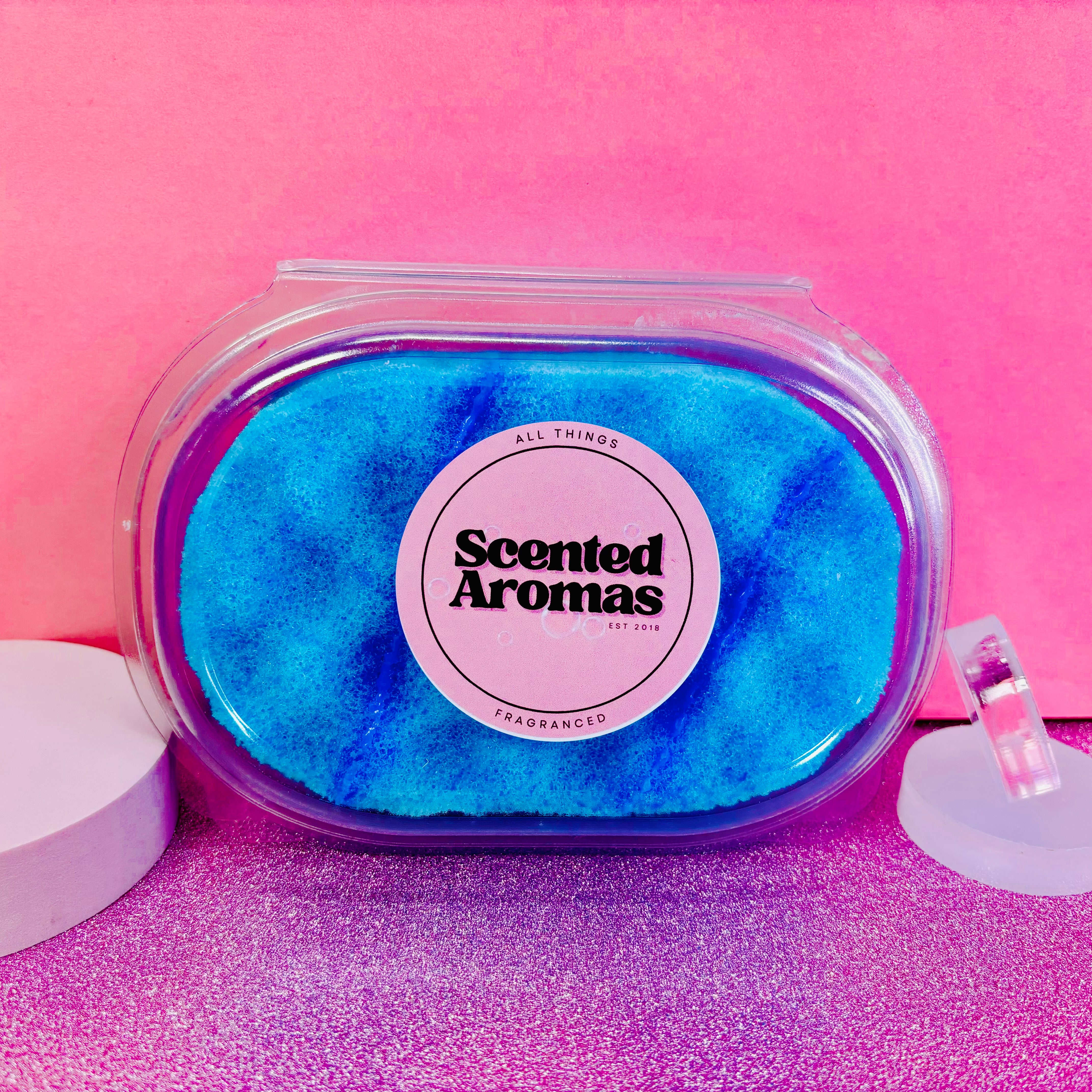 A pink labeled container showcasing the Bedtime Baby Bath Exfoliating Soap Sponge by The Soap Gal x, filled with soothing blue scented crystals, perfect for adding serenity to your skincare routine against a calming pink and purple backdrop.