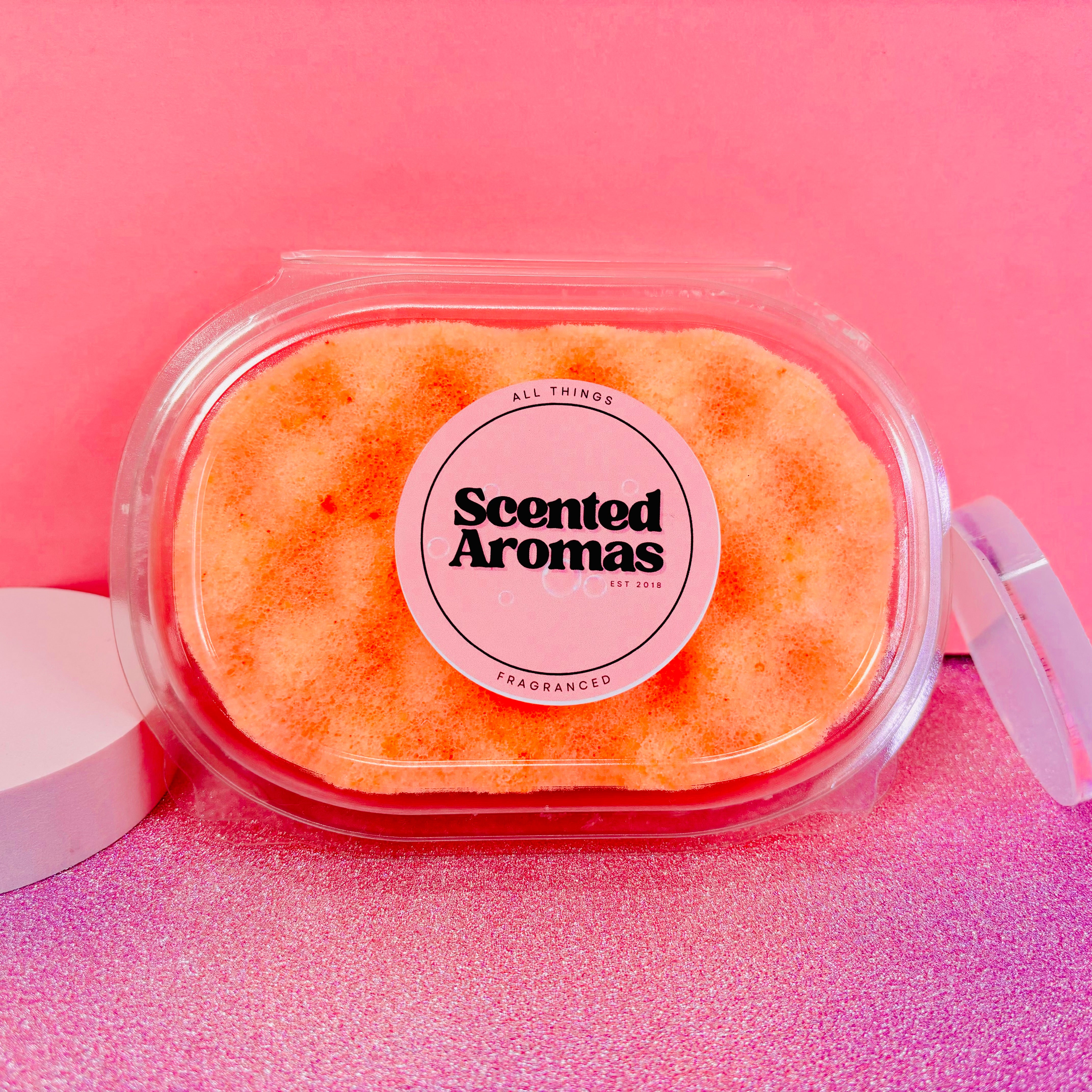 The Soap Gal x's Sweet Orange Exfoliating Soap Sponge, featuring a vibrant pink label, rests against a pink background, bringing a refreshing element to your skincare routine.