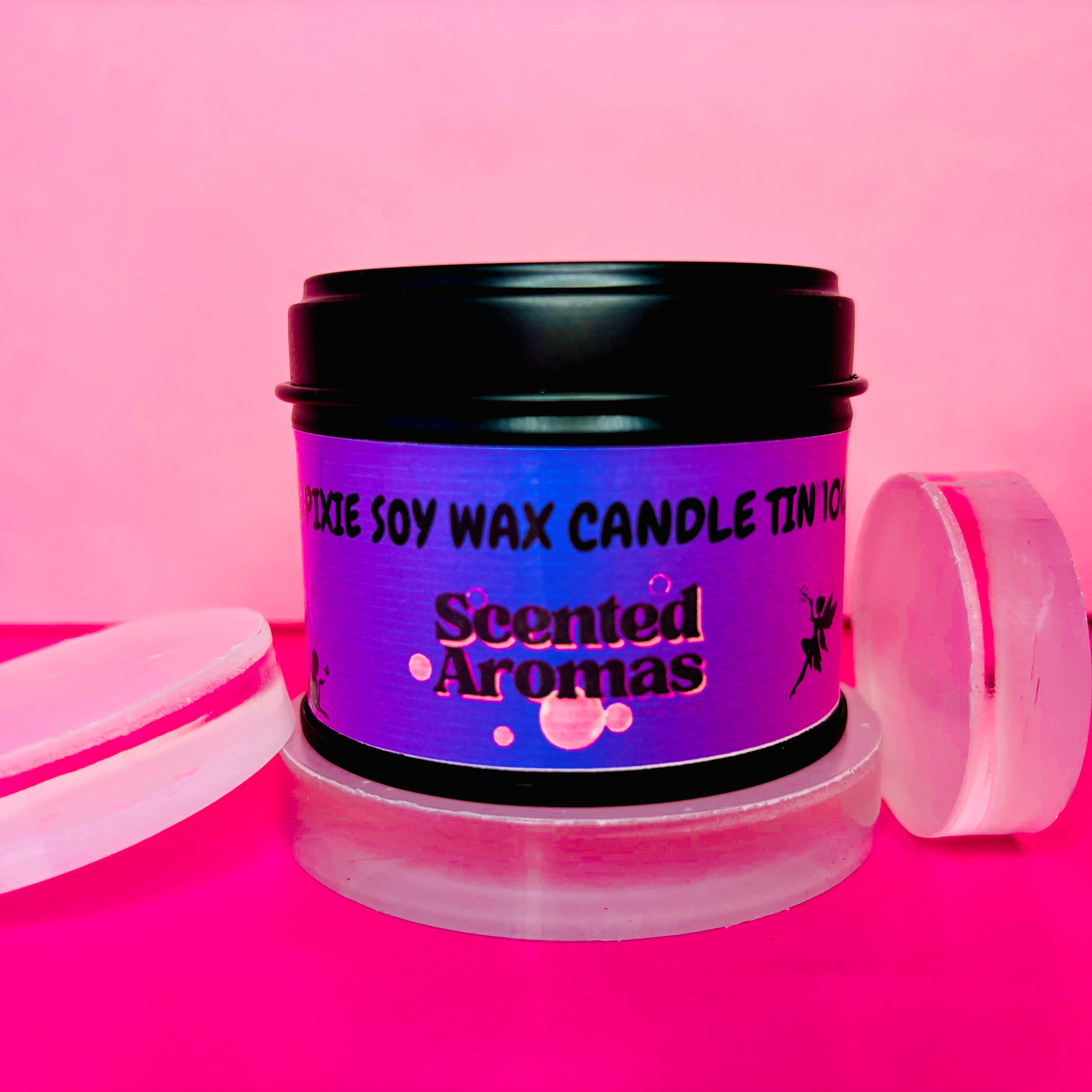 A purple tin labeled "Snow Pixie Candle Tin - 100ml" by The Soap Gal x contains a soy wax candle made from vegan-friendly ingredients, placed on two round glass slabs against a pink background. It's perfect for those who appreciate exceptional home fragrance experiences.