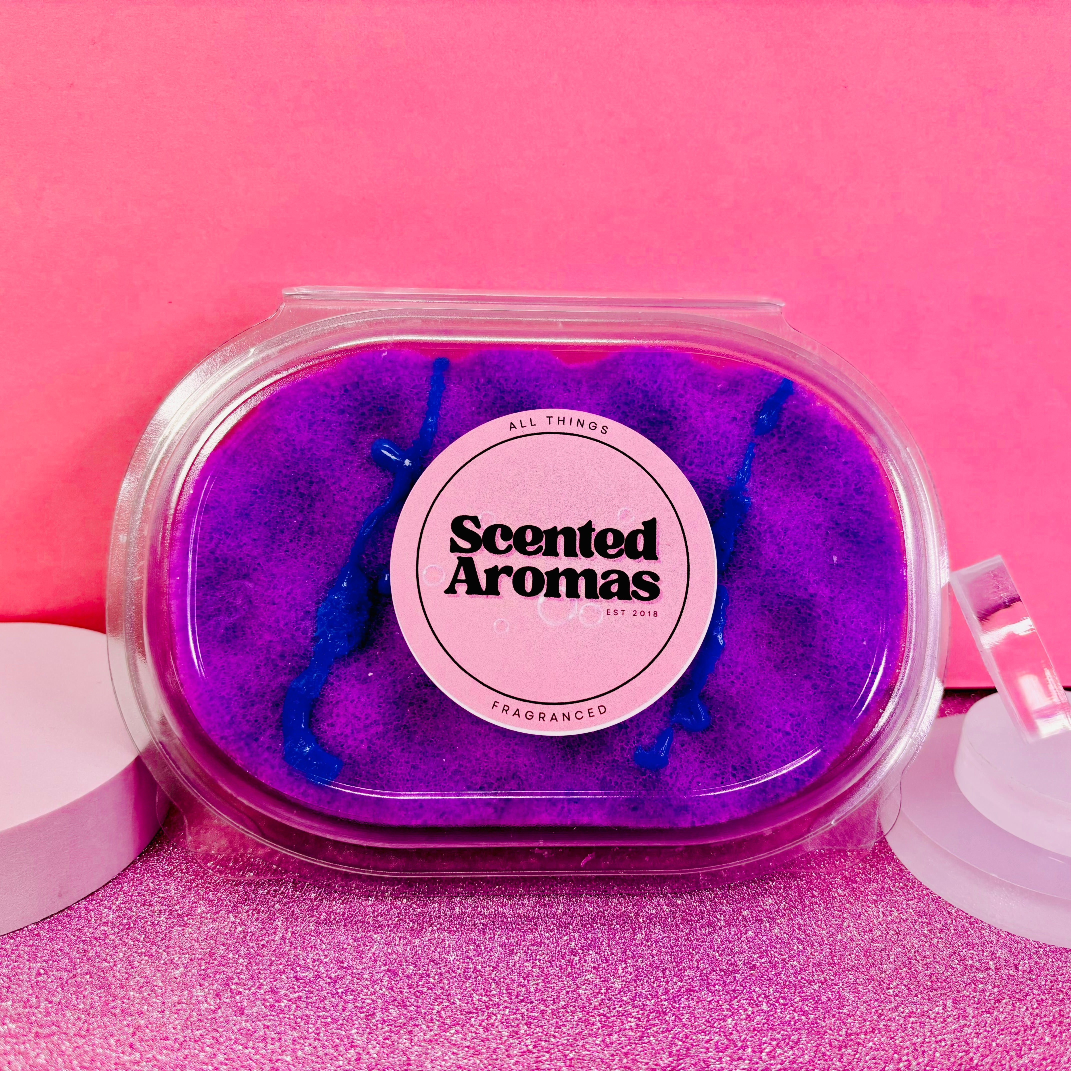 A transparent container holds purple scented wax labeled "Scented Aromas" set against a pink background, reminiscent of the Parma Violet Exfoliating Soap Sponge by The Soap Gal x.