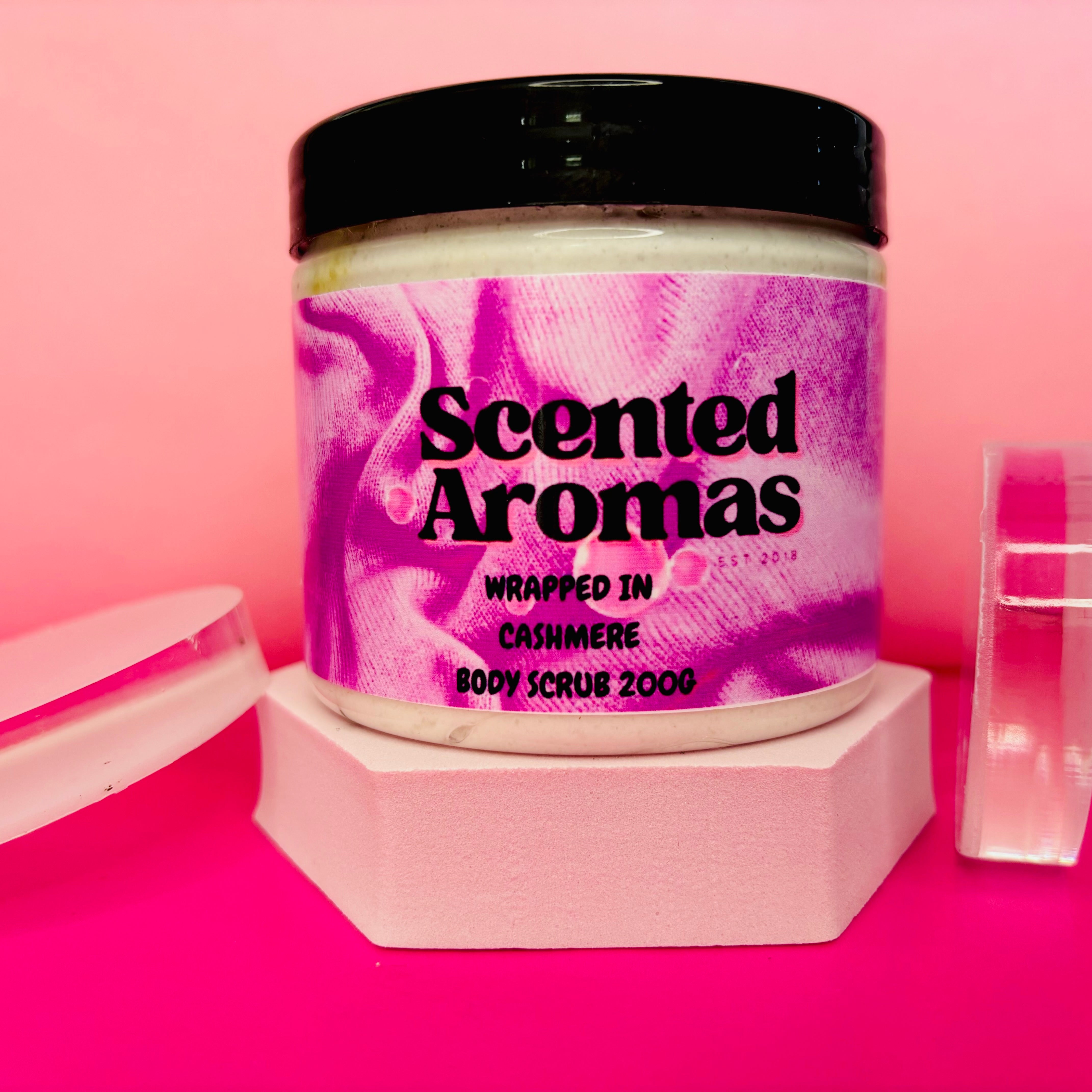 A jar of Wrapped in Cashmere Sugar Body Scrub 200g by The Soap Gal x, featuring a pink and purple design, elegantly perched on a pink pedestal.