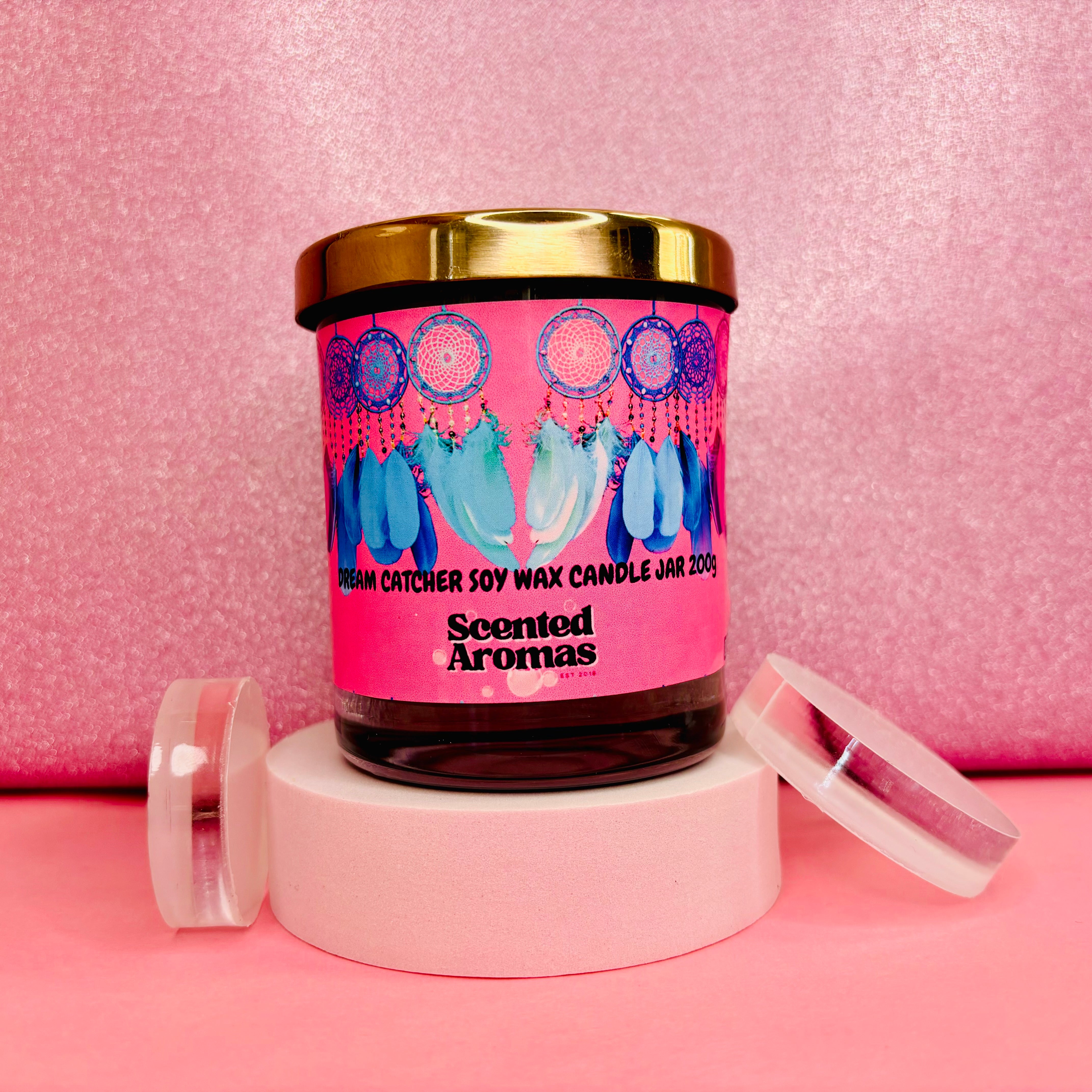 A jar candle from The Soap Gal x, labeled "Dream Catcher Candle - 200g," exudes a fruity floral scent, featuring a gold lid and a colorful dream catcher design on a pink background.