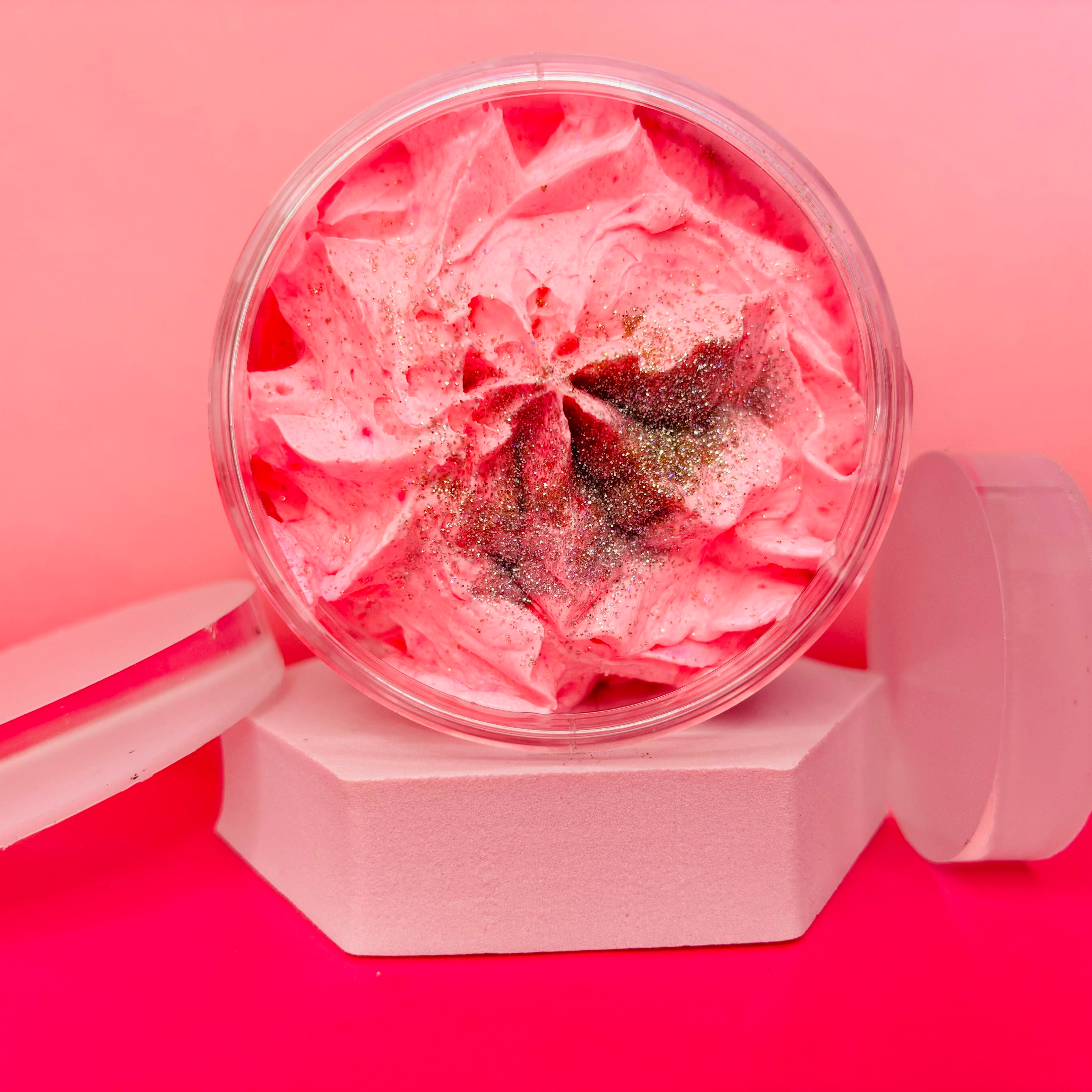 A 200g round container of The Soap Gal x Belle Of The Ball Sugar Body Scrub, featuring pink whipped cream subtly fragranced and topped with glitter, rests elegantly on a white hexagonal stand against a pink background.