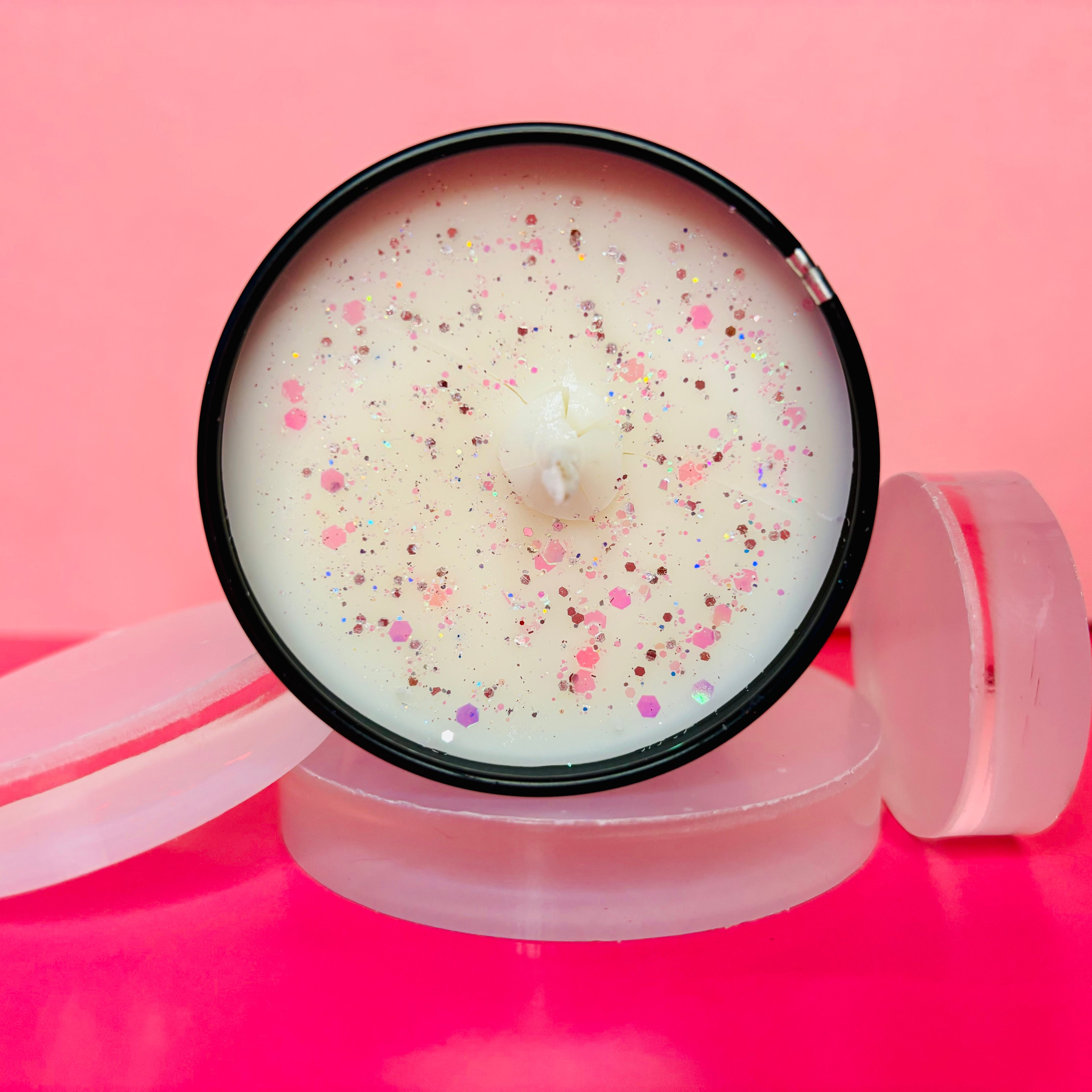 An overhead shot of The Soap Gal x's Apple Candy Candle Tin - 100ml, crafted from soy wax with a hint of glitter, housed in a sleek black container and set on frosted circular discs against a pink backdrop.