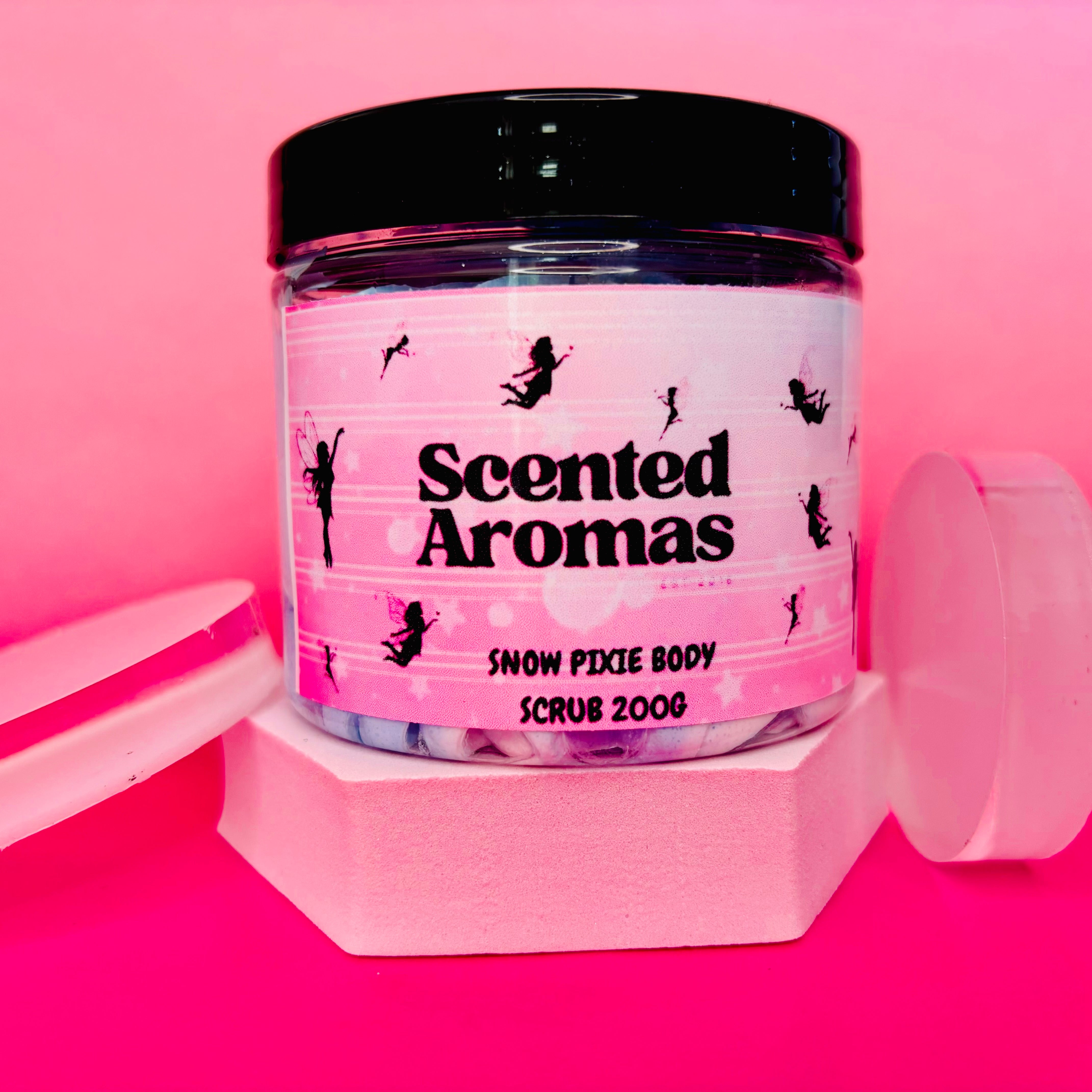 A jar of "The Soap Gal x" Snow Pixie Sugar Body Scrub 200g sits against a pink background. This vegan, exfoliating scrub showcases a label adorned with silhouette figures.