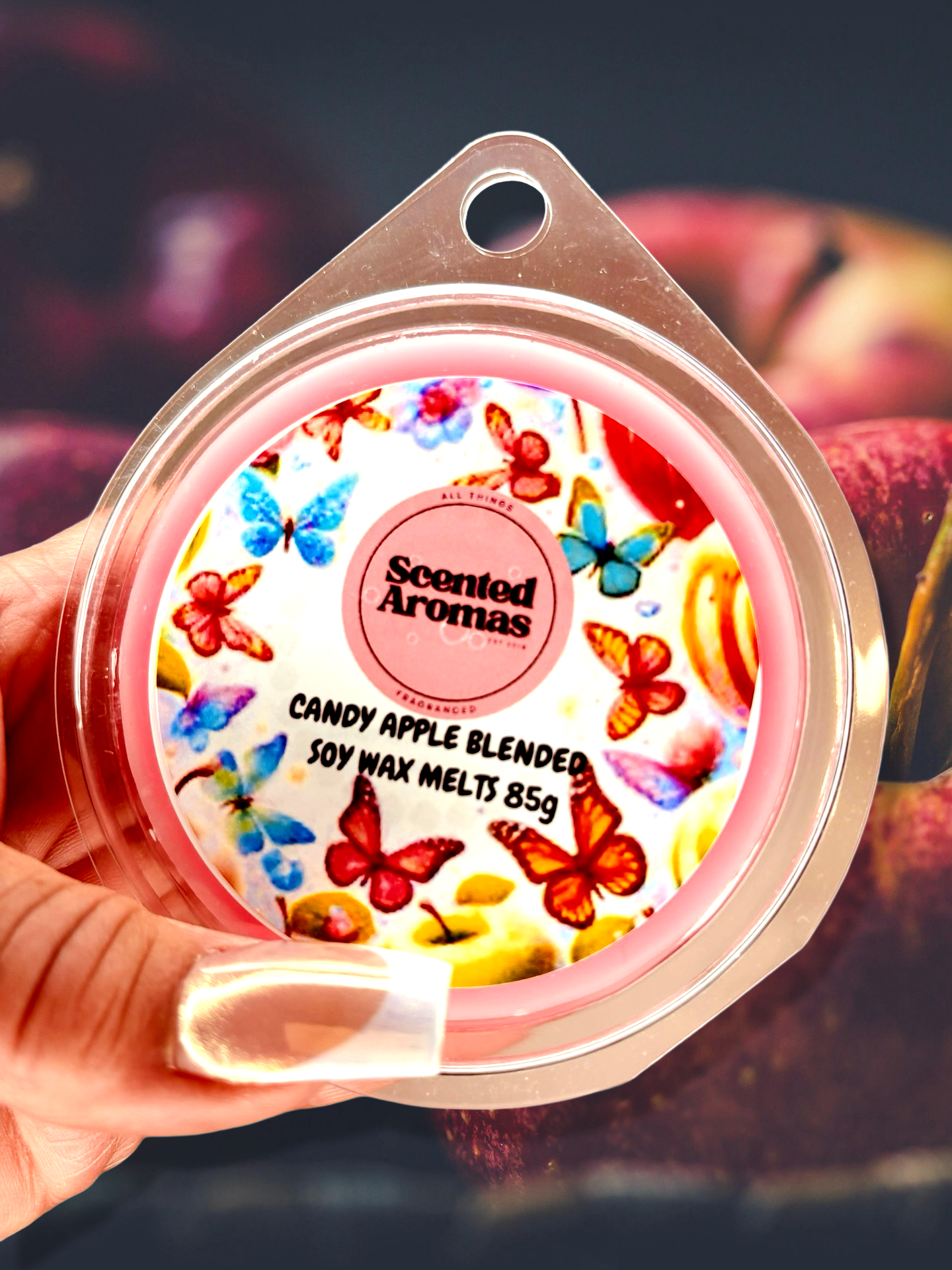 A hand holds a package of The Soap Gal x Candy Apple Wax Melts 85g, showcasing butterfly designs on the lid. These wax melts promise to fill your space with unique scents, enhancing the ambiance in any room.