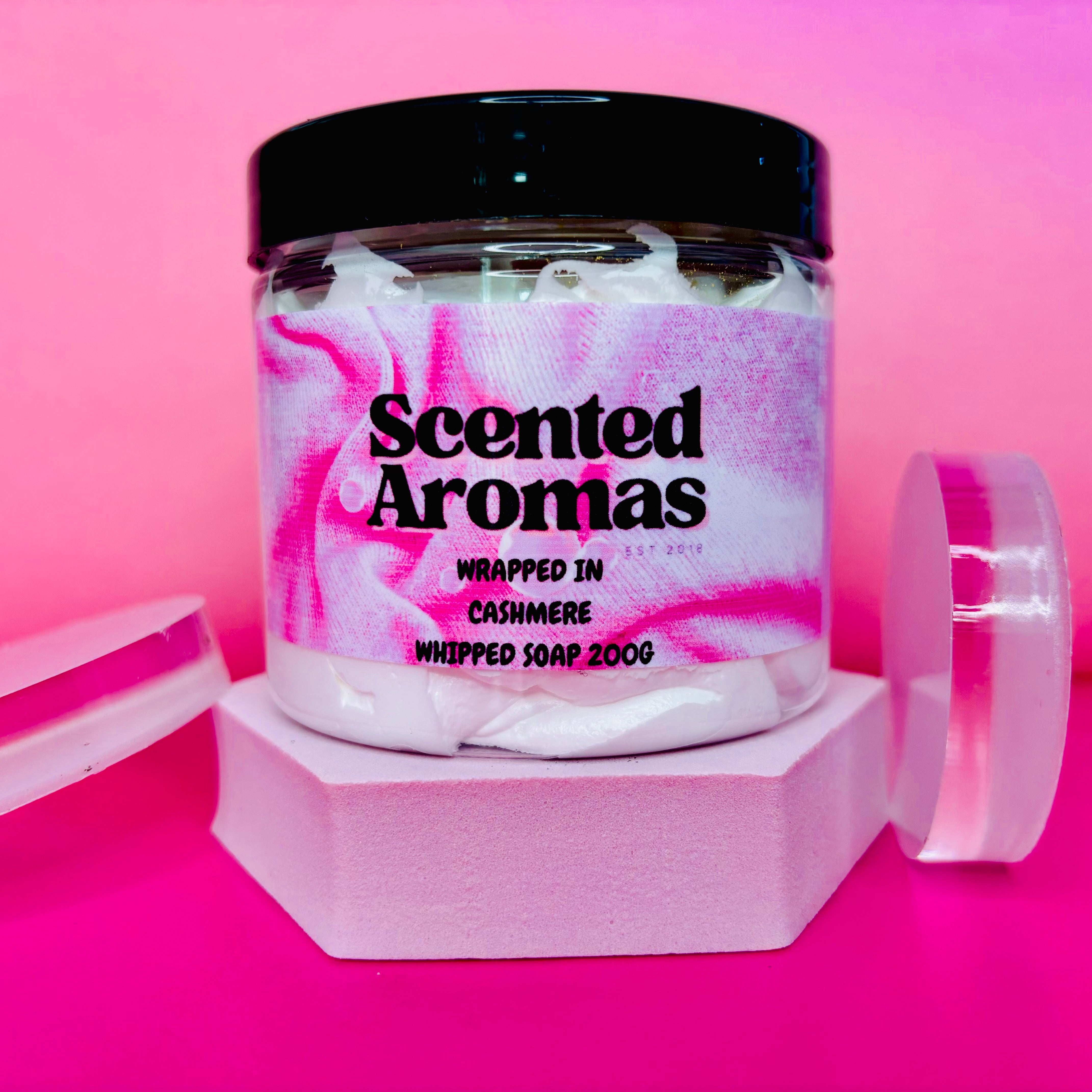 A 200g jar of "Wrapped in Cashmere" Whipped Soap Shave Butter by The Soap Gal x, known for its sweet musk fragrance, is displayed elegantly on a pink background.