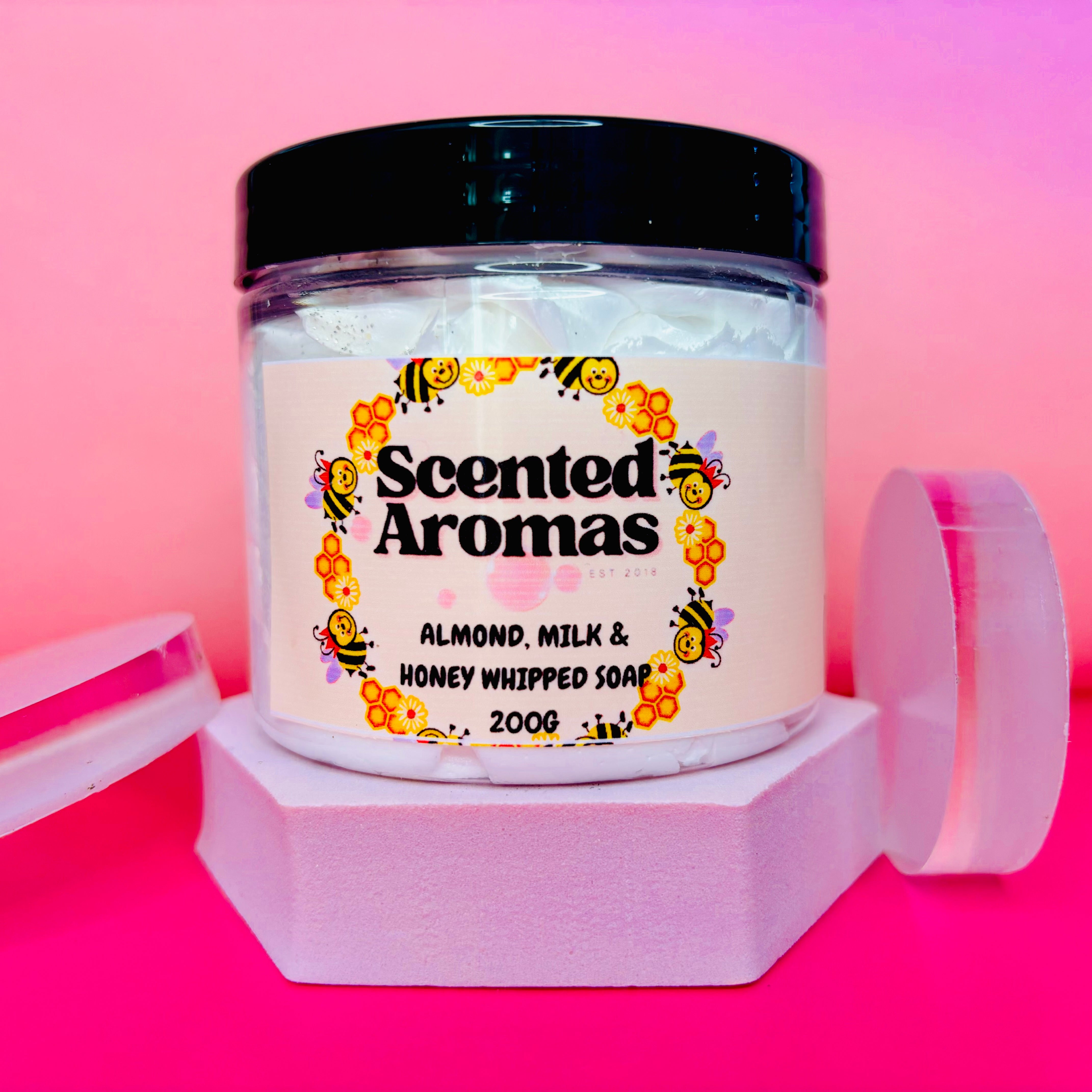 A jar of The Soap Gal x Almond Milk and Honey Whipped Soap Shave Butter 200g is elegantly positioned on a hexagonal base against a pink gradient background, promising a luxurious solution for strawberry legs.