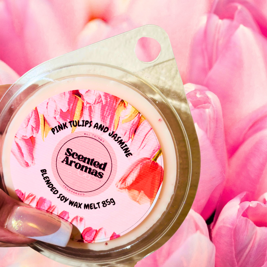 A hand delicately holds "The Soap Gal x" vegan soy wax melt labeled "Pink Tulip and Jasmine Wax Melts 85g," set against a charming backdrop of vibrant pink tulips.