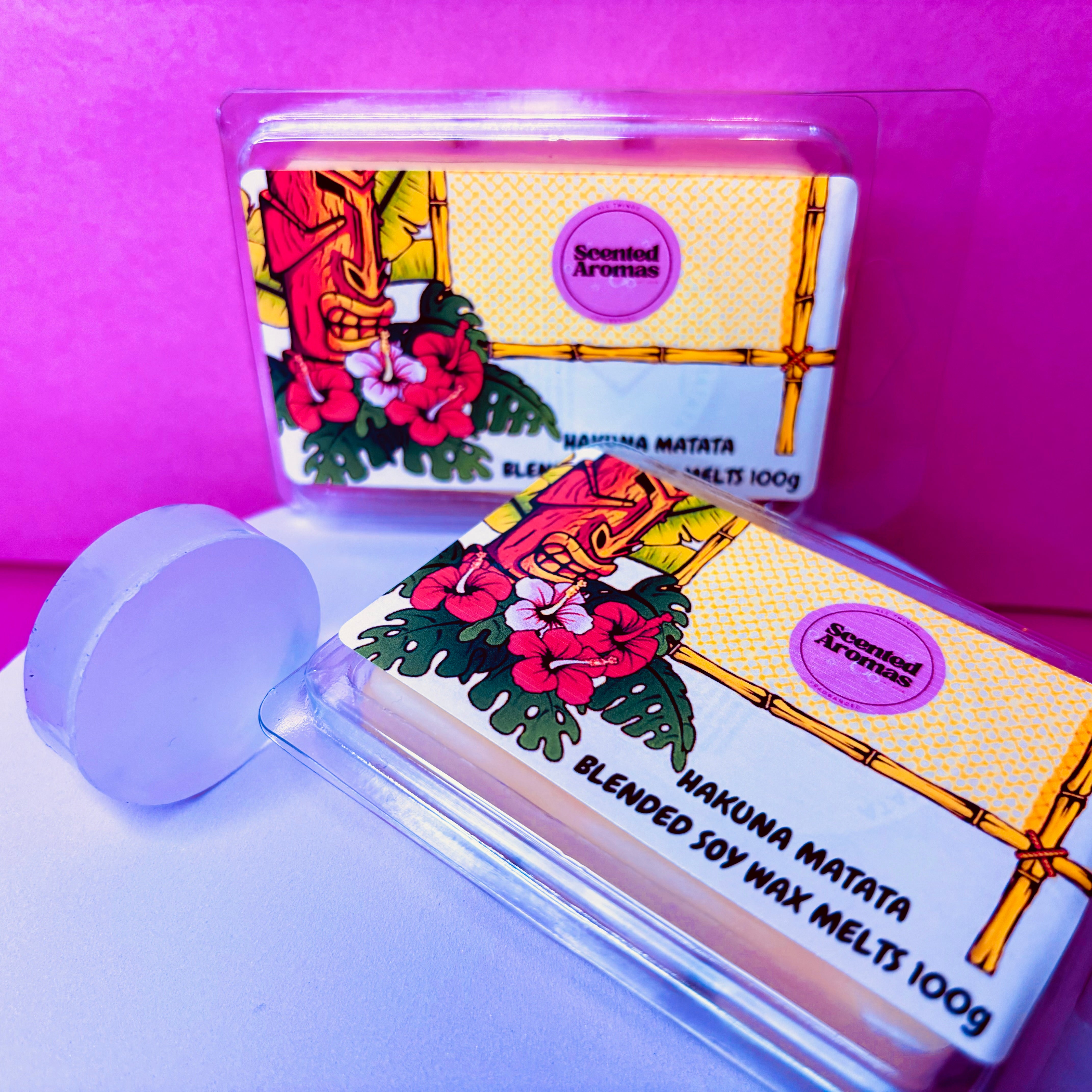 Two packages of "It Means No Worries" wax melts, weighing 85g each and crafted by The Soap Gal x, are showcased against a pink backdrop. These blended soy wax melts feature citrus scents complemented by floral and tiki designs. Made from biodegradable wax, they provide an eco-friendly way to enjoy delightful fragrances.