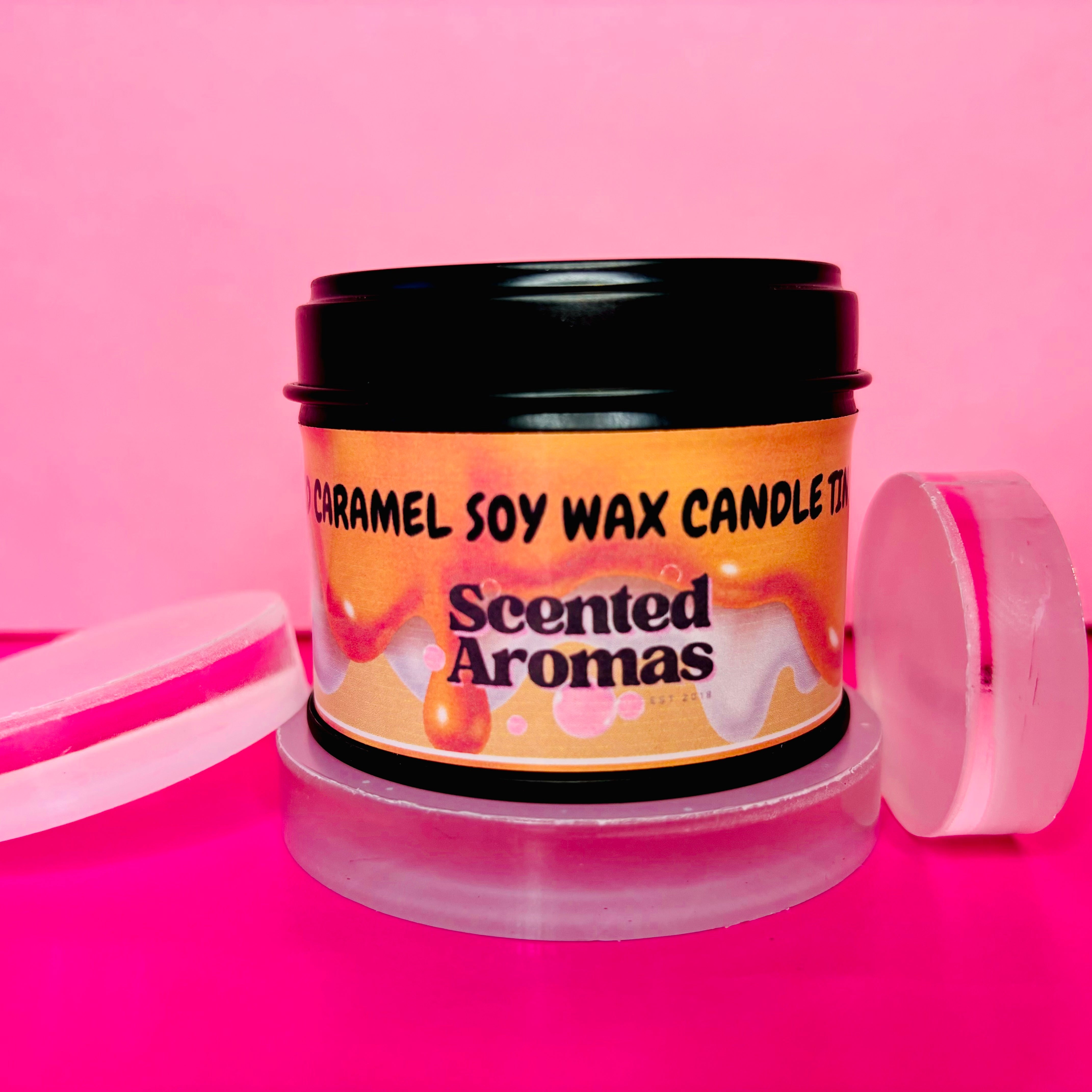 The Salted Caramel Candle Tin - 100ml from "The Soap Gal x" offers a long-lasting scent, with its soy wax composition providing hours of aromatic pleasure. Presented with its lid off against a vibrant pink background, it serves as both a sensory delight and an elegant decor item.