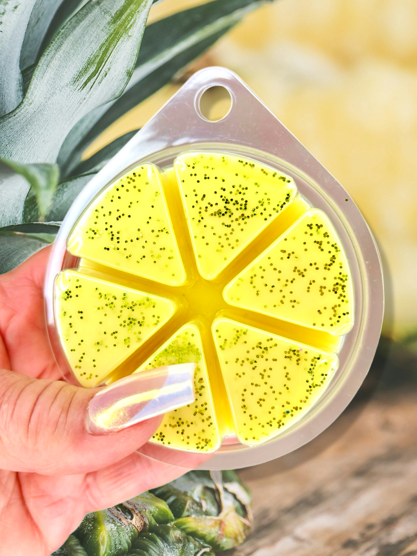 A hand holds a segmented circular container with yellow gel and small black specks, resembling a lemon slice, evoking a fruity atmosphere. The container is filled with Pineapple Whip Wax Melts 85g by The Soap Gal x.