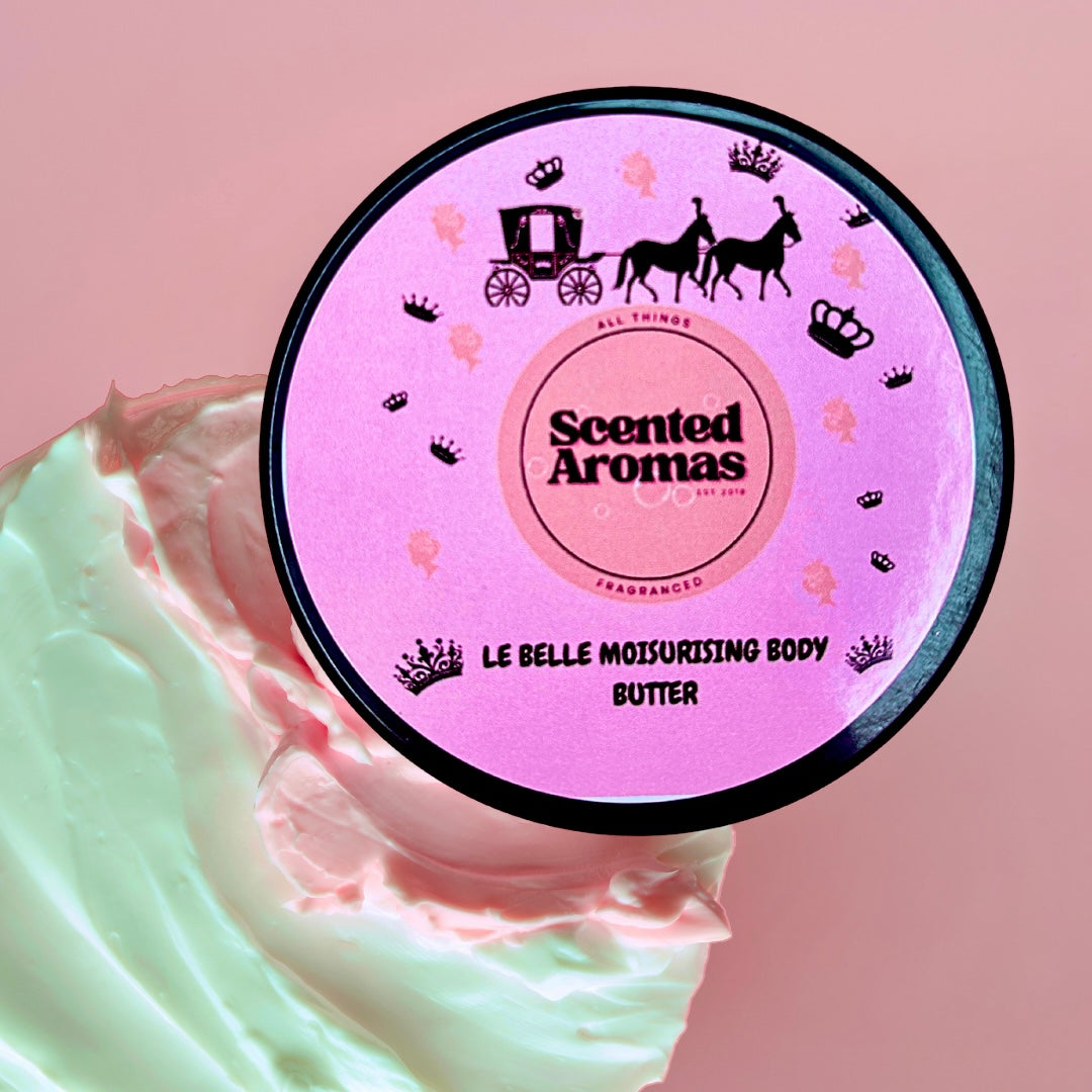 A round container of Belle Of The Ball Moisturising Body Butter 100ml from The Soap Gal x is displayed on a swirl of cream against a pink background. The label, decorated with a horse-drawn carriage and crowns, suggests a floral and leather fragrance with a nourishing formula.