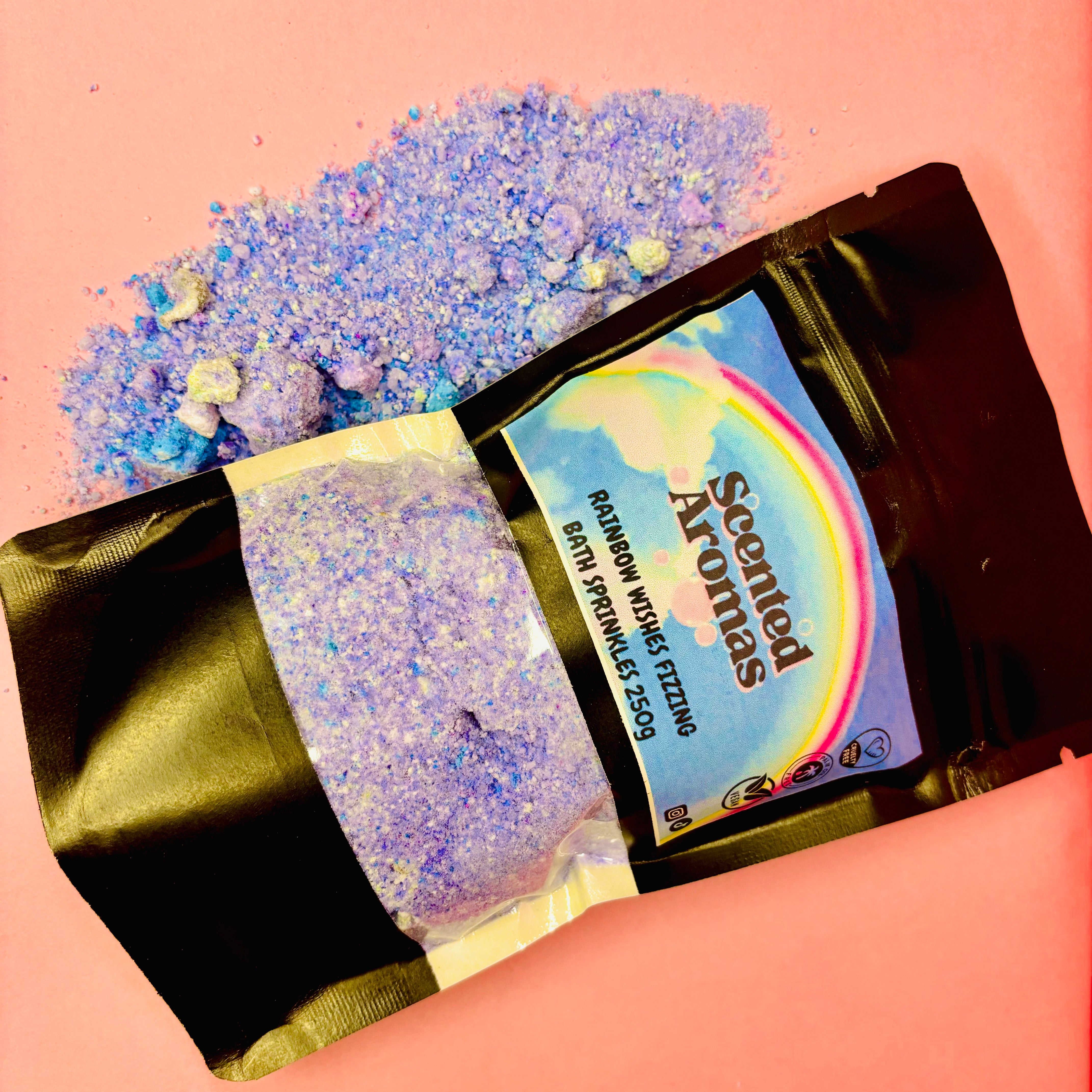 A black pouch of blue and purple bath salts, featuring Epsom salts, is spilled on a pink surface. The label reads "Rainbow Wishes Bath Fizzing Salts 250g." from The Soap Gal x.