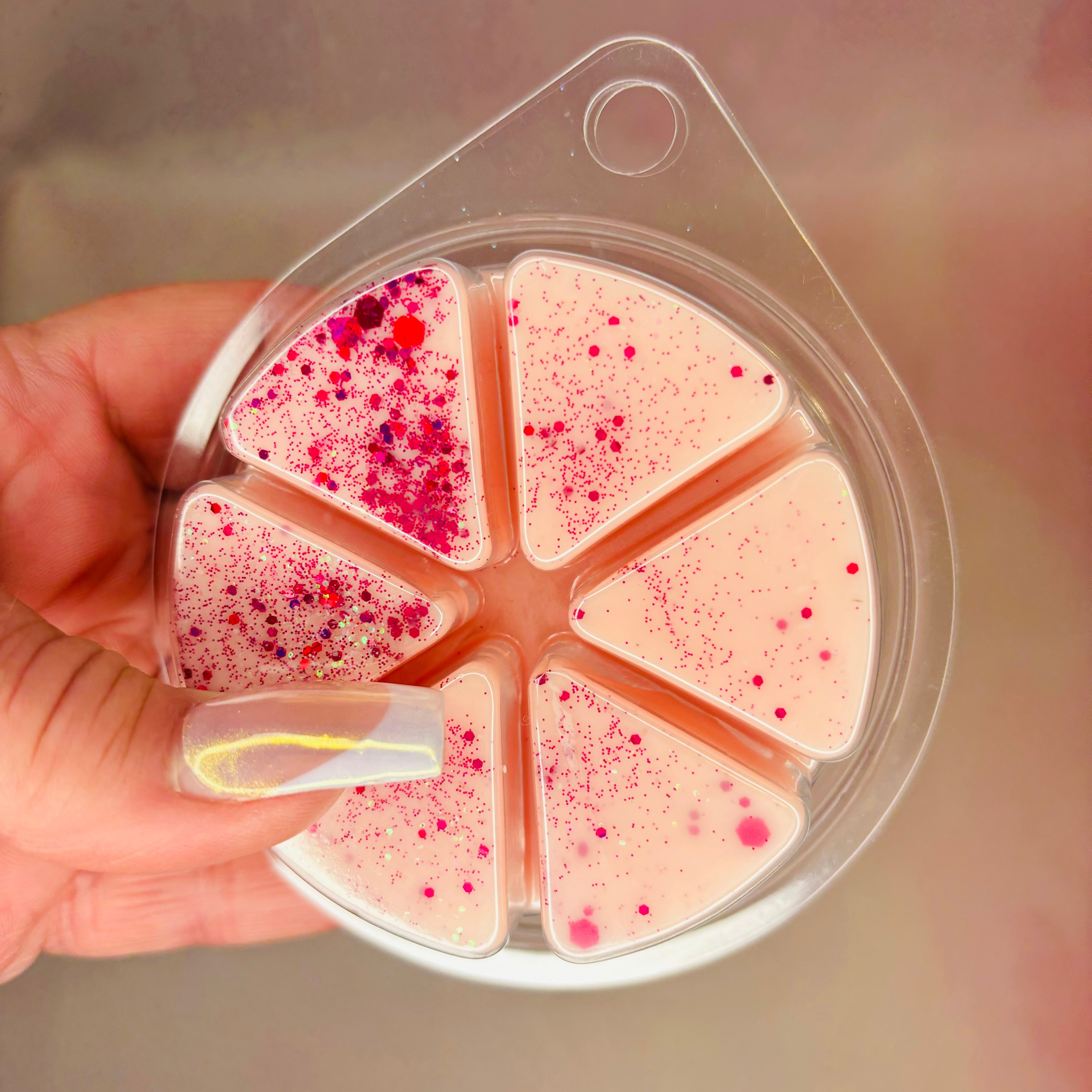 A hand holds a circular container featuring six pink triangular pieces, adorned with red speckles and glitter, unveiling the captivating Rhubarb and Rose Wax Melt 85g by The Soap Gal x. Made from a soy wax blend, these melts ensure a long-lasting aroma that transforms your space into a fragrant sanctuary.