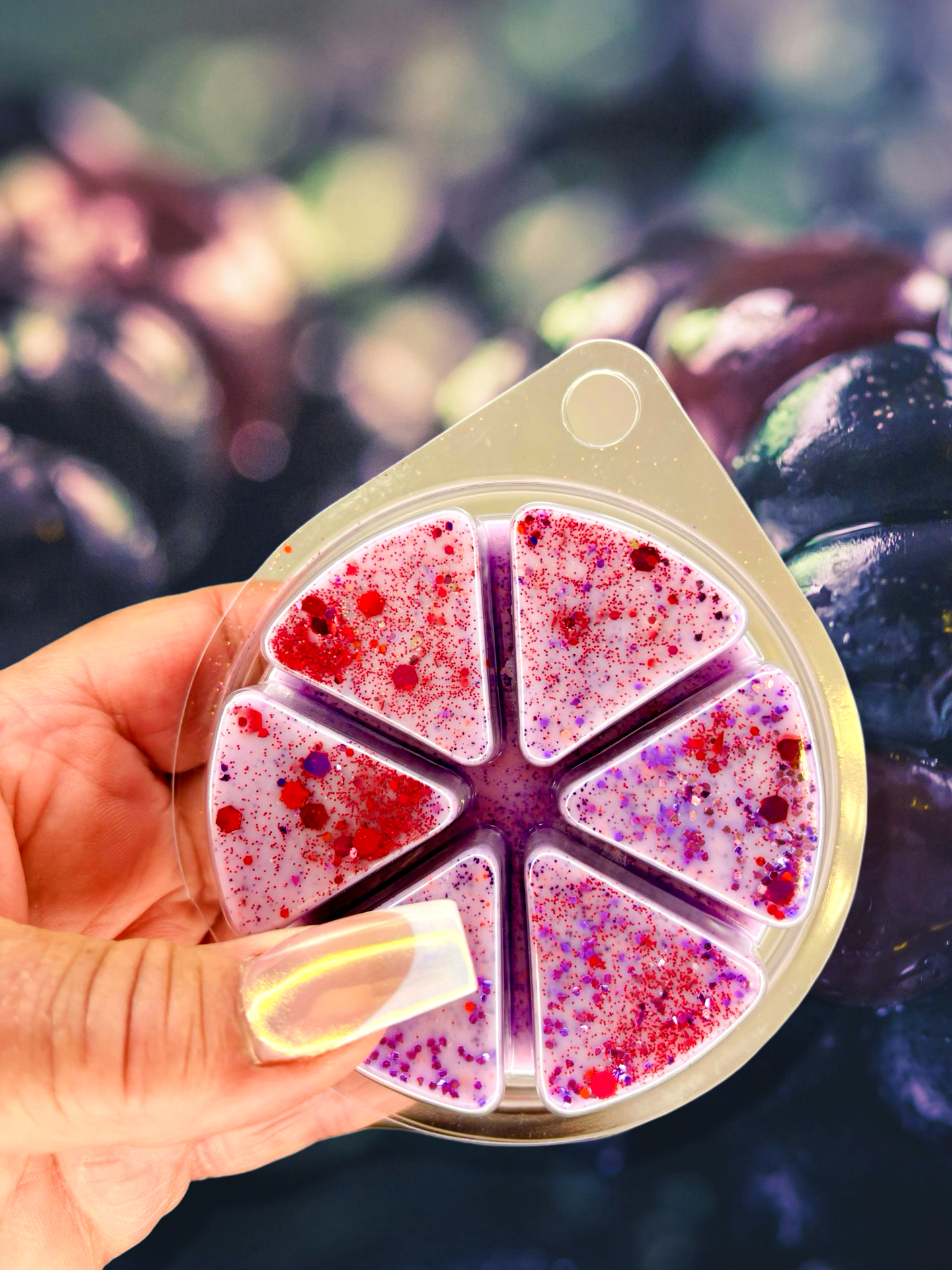 A hand holds a circular container with six triangular segments of purple and red speckled material, similar to The Soap Gal x's Blackberry Cider Wax Melt 85g. In the background, dark berries blur softly—a nod to its handmade Soy Wax blend origin in the UK.