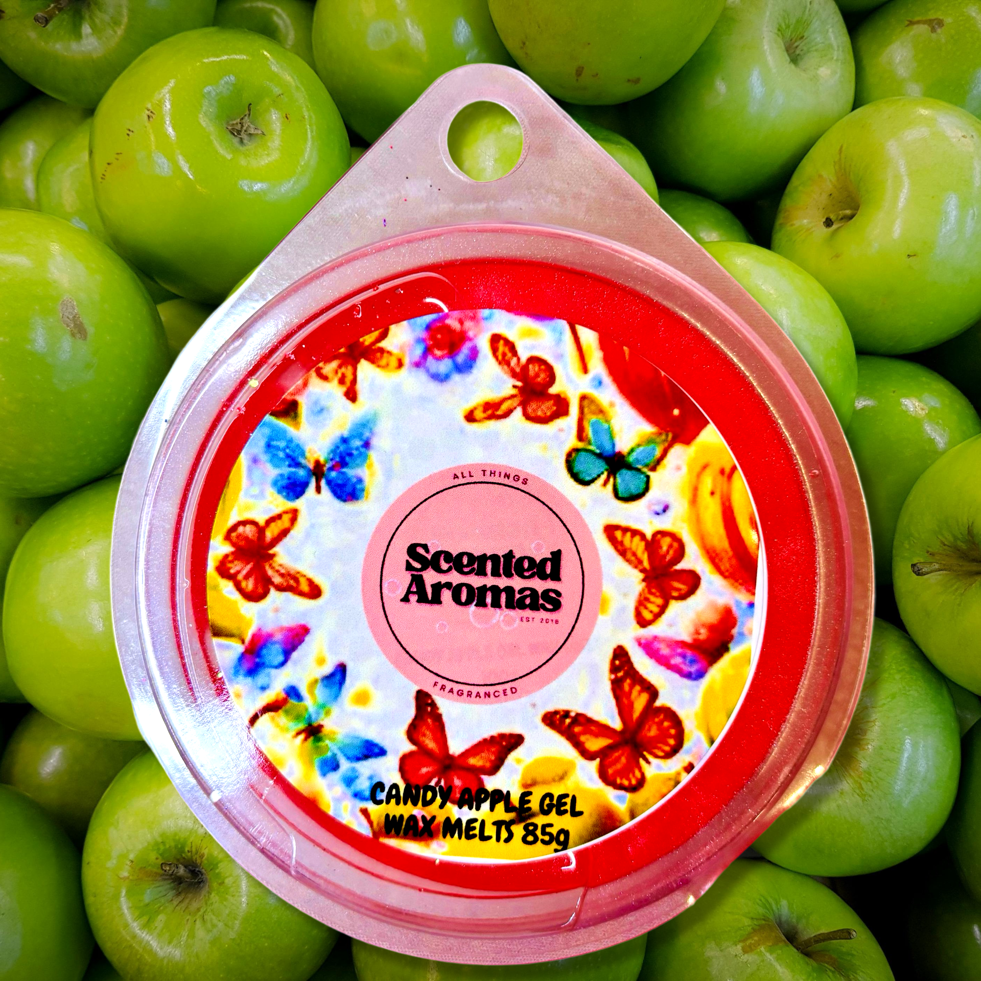 An 85g round container of "Frosted Candy Apple Gel Wax Melts" by The Soap Gal x rests on green apples, with its lid featuring vibrant butterfly illustrations that encapsulate the essence of Scented Aromas.