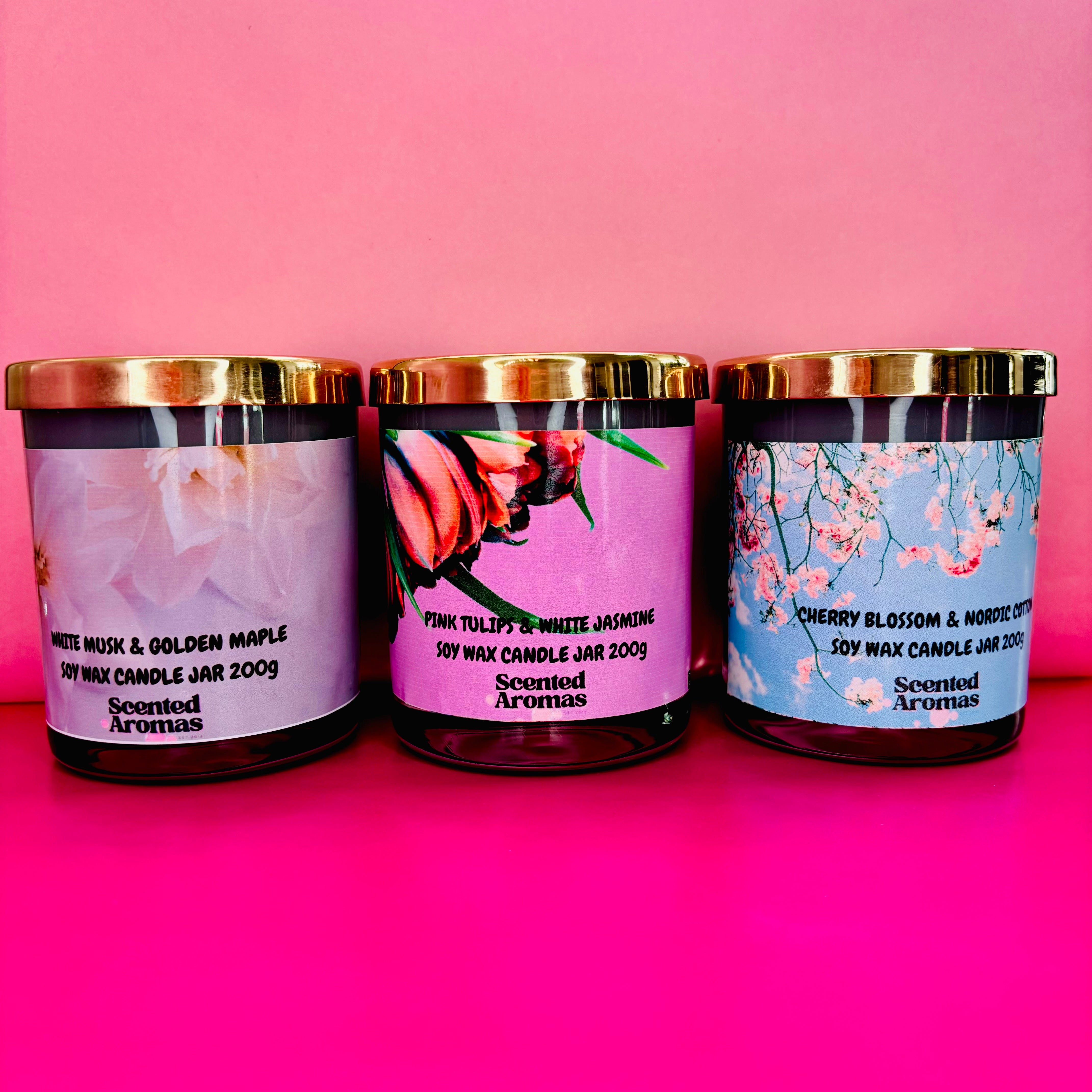 Three scented candles, including "Cherry Blossom and Nordic Cotton" by The Soap Gal x, with enticing labels like "White Musk & Golden Maple" and "Pink Tulips & White Jasmine," create inviting aromas against a pink background.