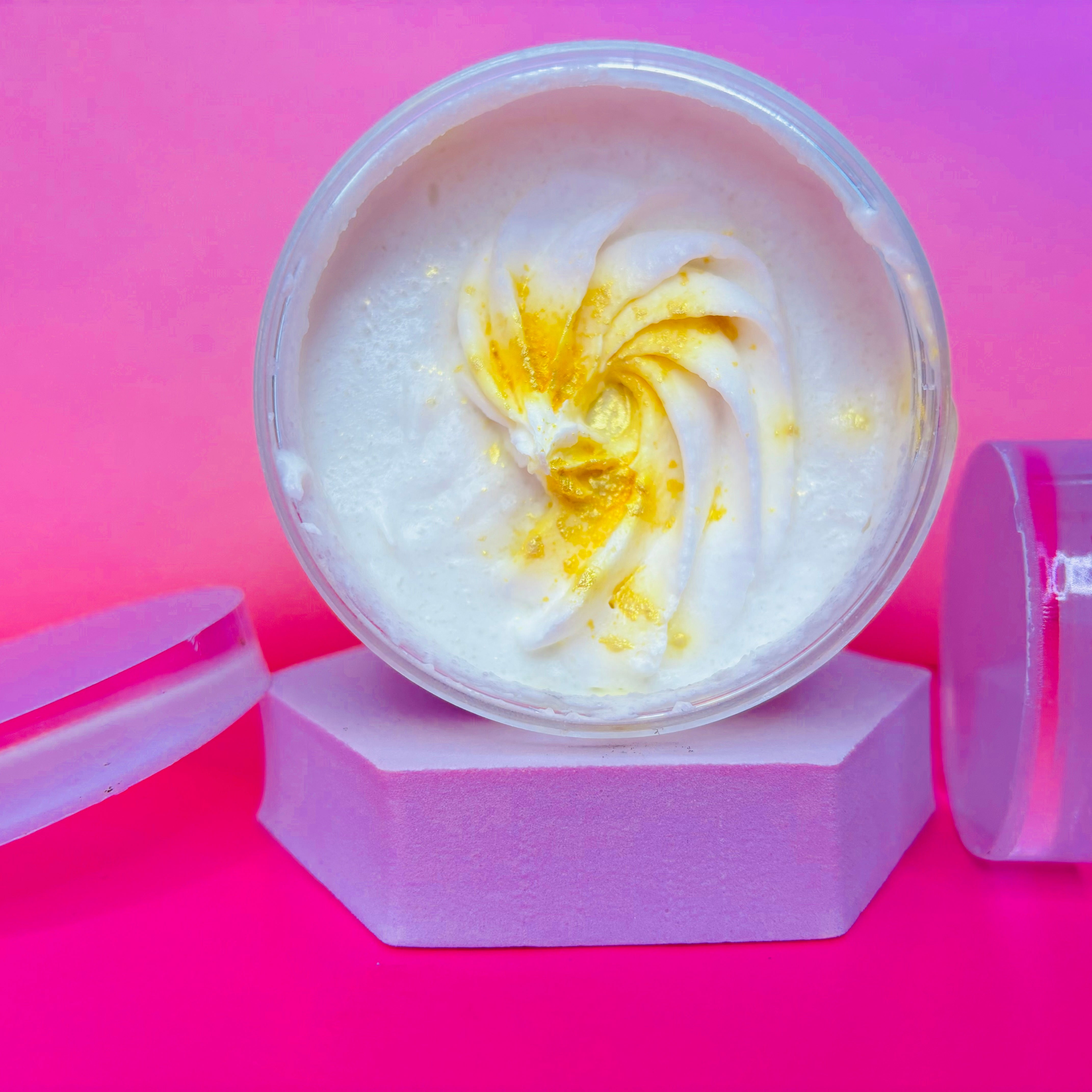 Open jar of The Soap Gal x Wrapped in Cashmere Sugar Body Scrub, featuring a swirl and gold accents, placed on a geometric stand against a pink and purple gradient background.