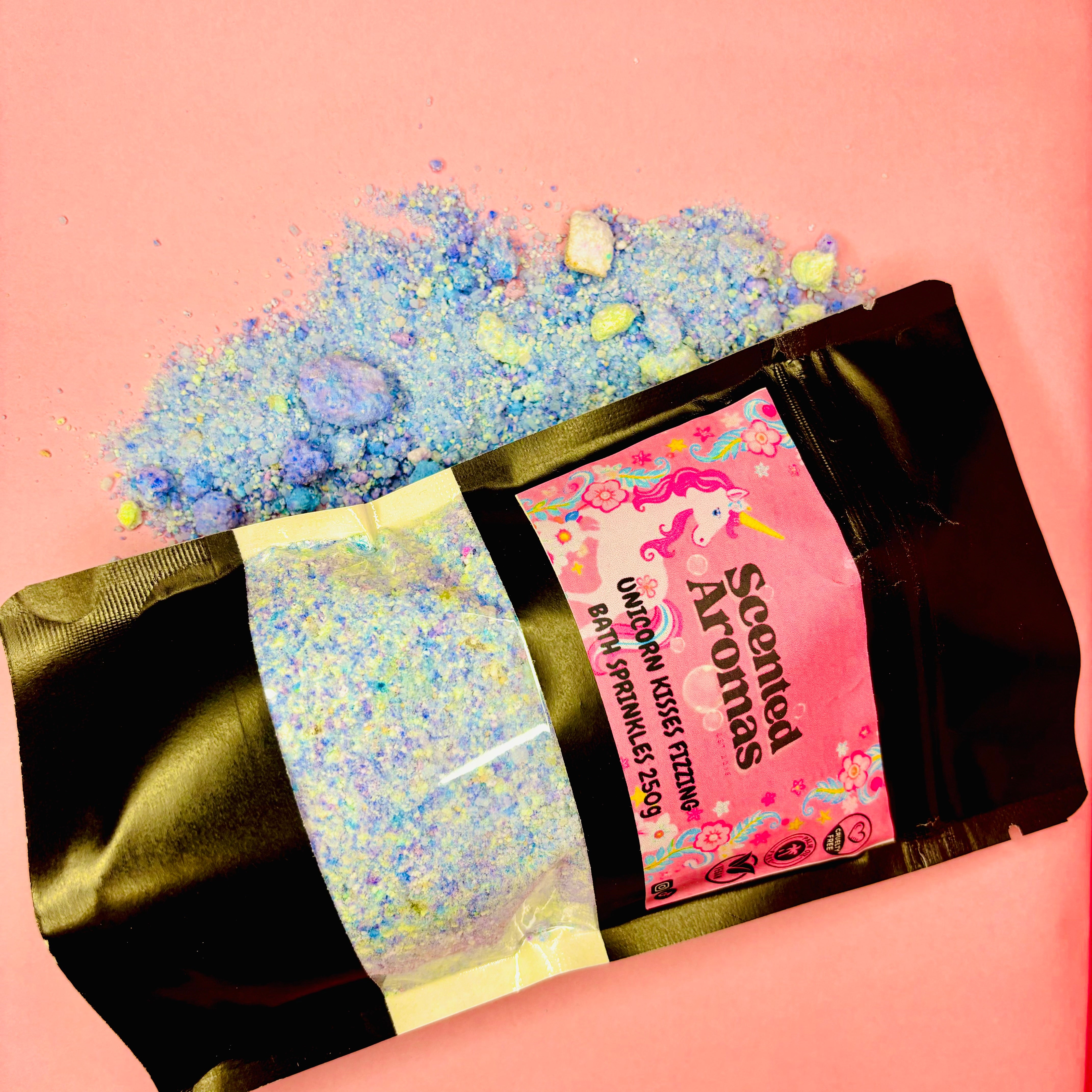 A pink bag filled with The Soap Gal x's Unicorn Kisses Bath Fizzing Salts releases its fruity floral aroma onto a pink surface. The label reads "The Soap Gal x Unicorn Kisses Bath Fizzing Salts, 250g.