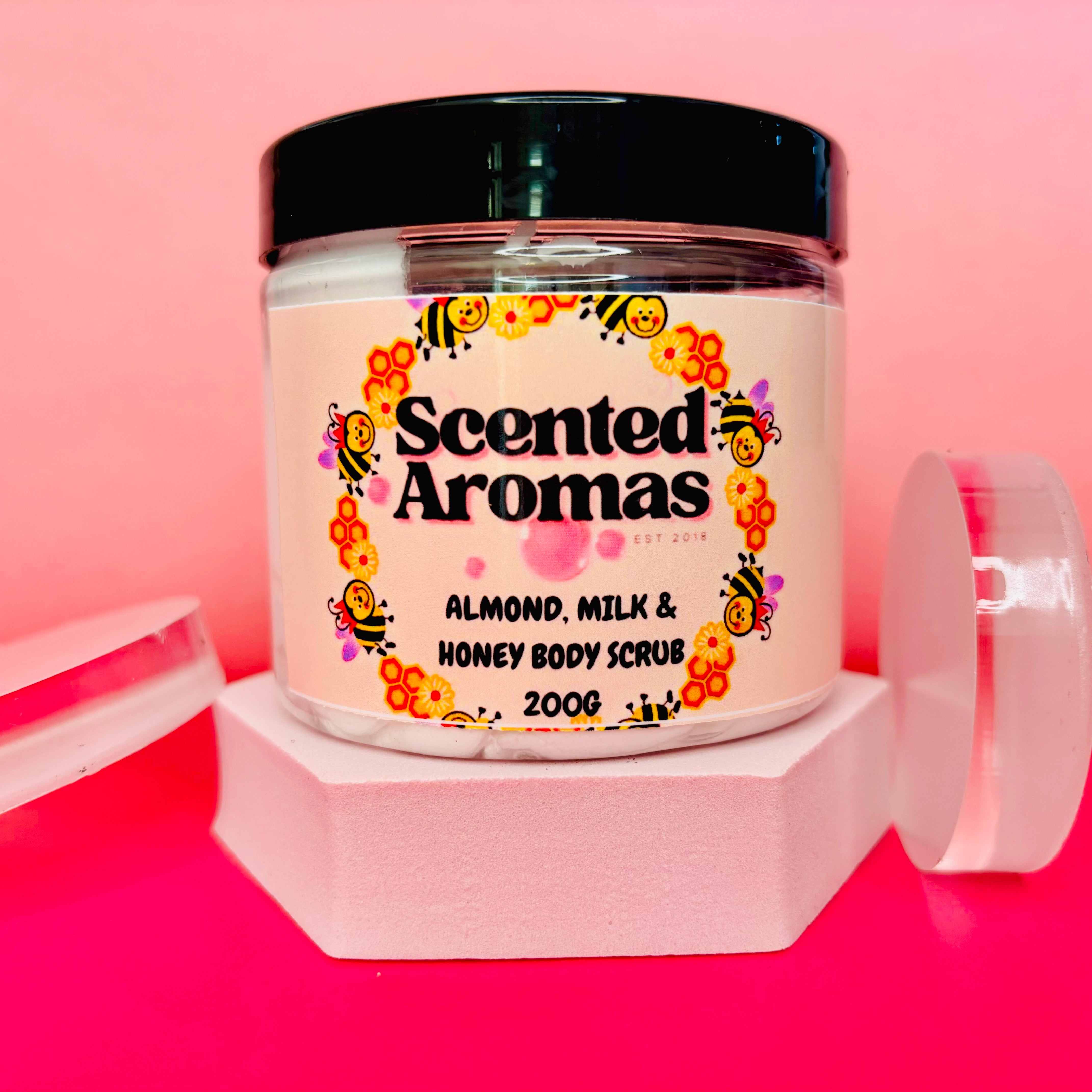 A "Scented Aromas" Almond Milk and Honey Sugar Body Scrub 200g sits on a pink background on a hexagonal white stand, promising smooth, glowing skin.