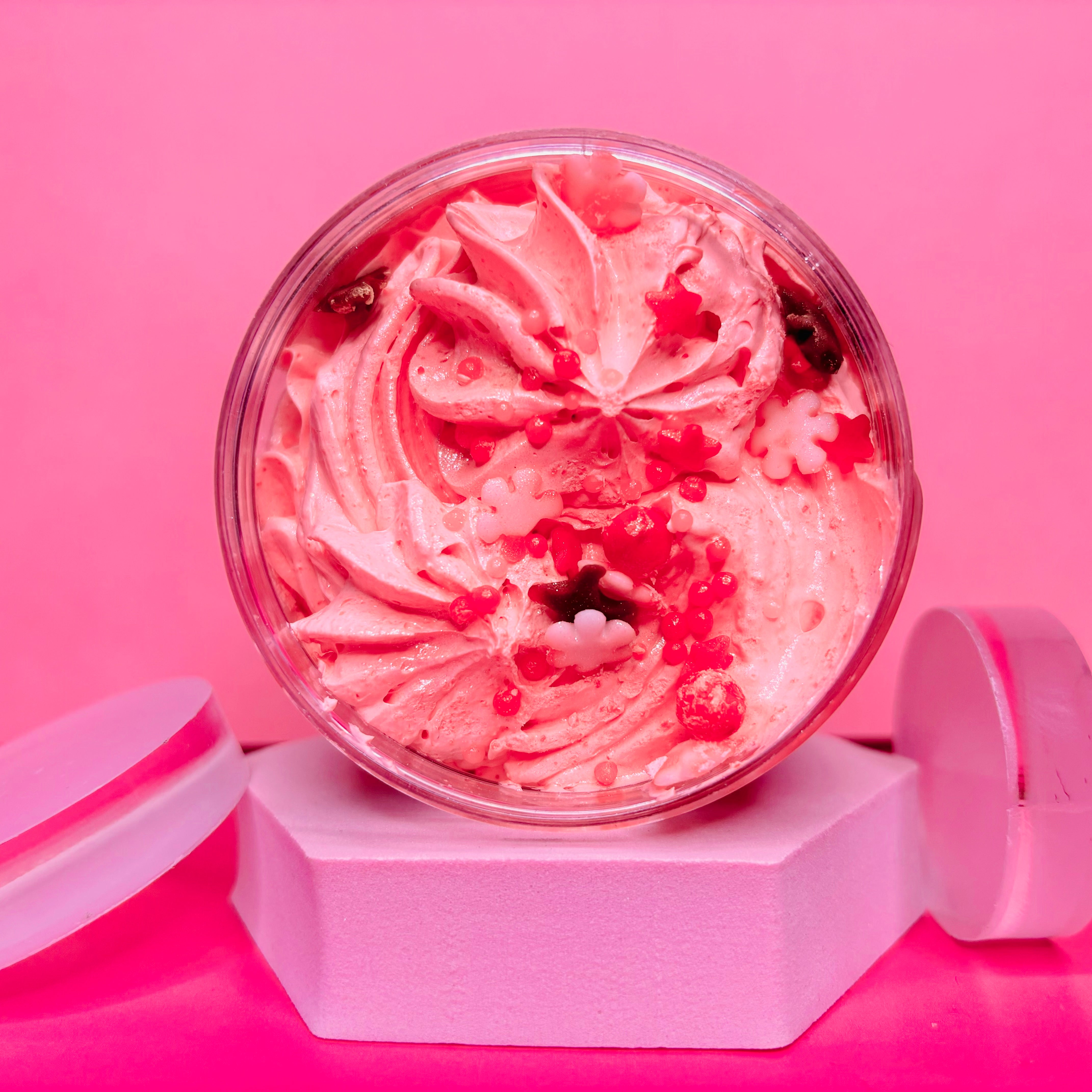 A jar of The Soap Gal x Christmas Cookies Whipped Soap Shave Butter, adorned with decorative sprinkles and flower shapes, is elegantly displayed on a pink hexagonal pedestal against a matching pink background, evoking the charm of festive Christmas cookies.