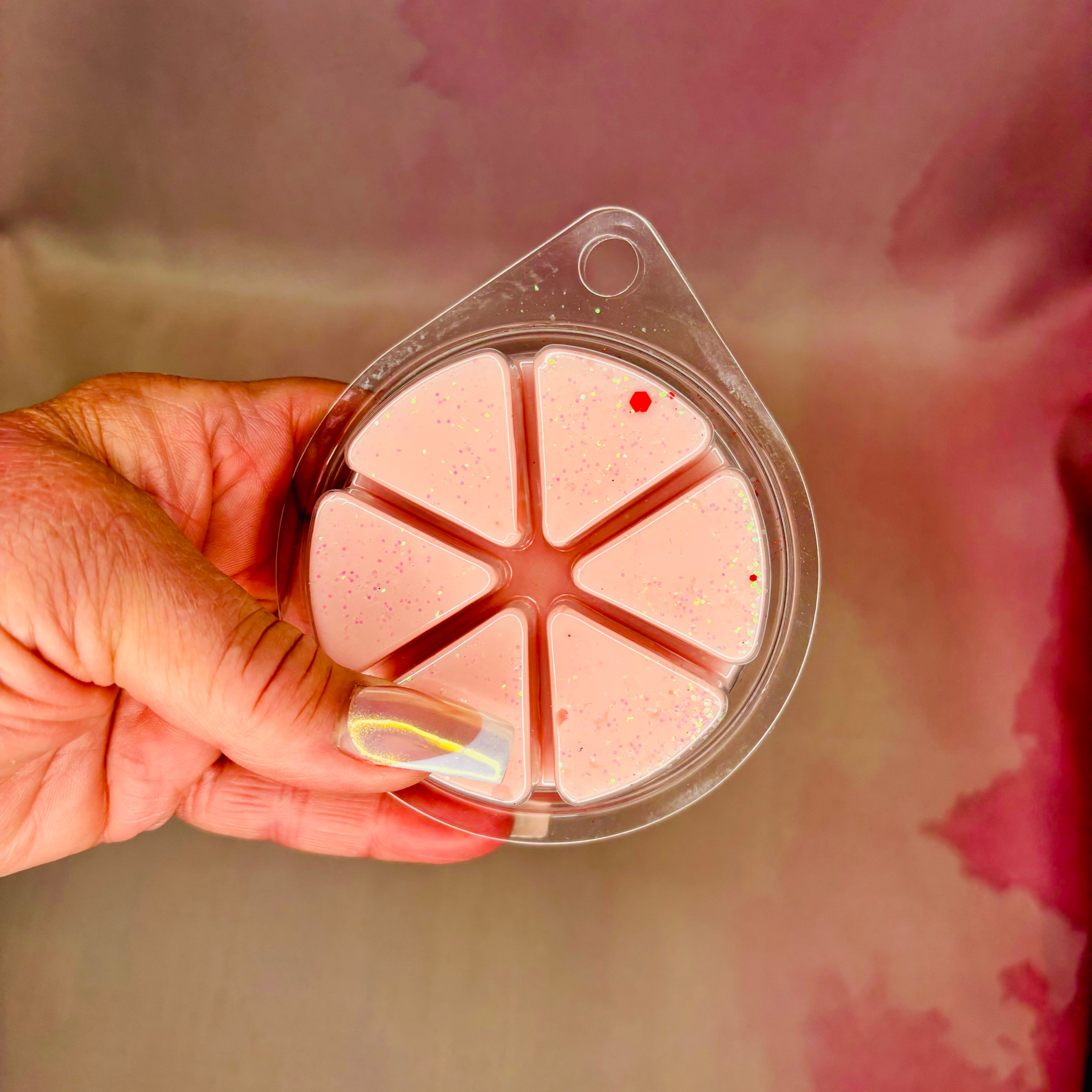 A hand holds a round clear plastic container with six pink, glittery, triangular soy wax melts on a pink background. These "Home Sweet Home Wax Melt | 85g" by The Soap Gal x are perfect for enhancing your home fragrance experience.