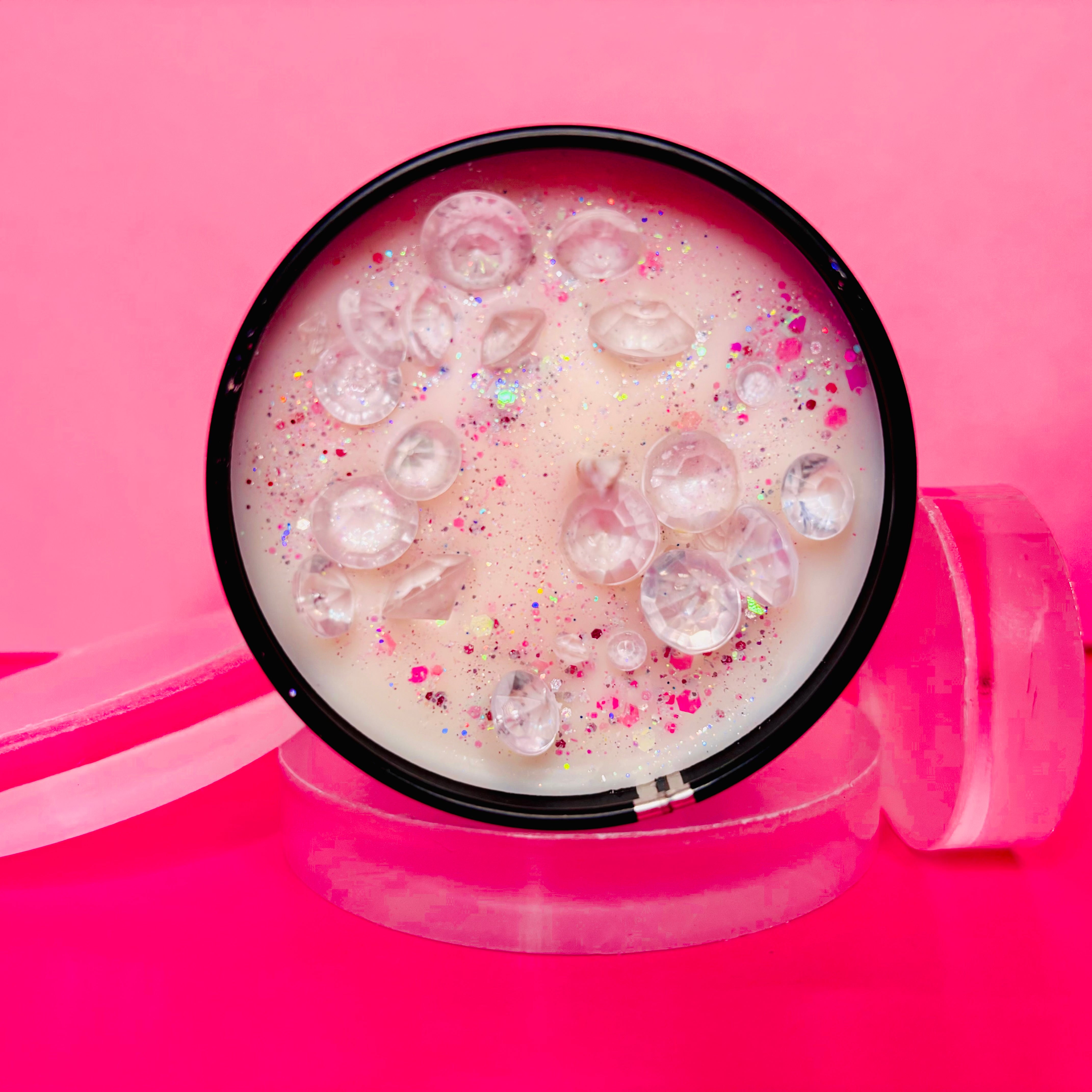 A round container, akin to The Soap Gal x Ombre Nomade Candle Tin - 100ml, is filled with a pink and white creamy substance and adorned with clear bubbles and glitter. Set against a pink background featuring translucent circular elements, it exudes the allure of luxury woody fragrance.