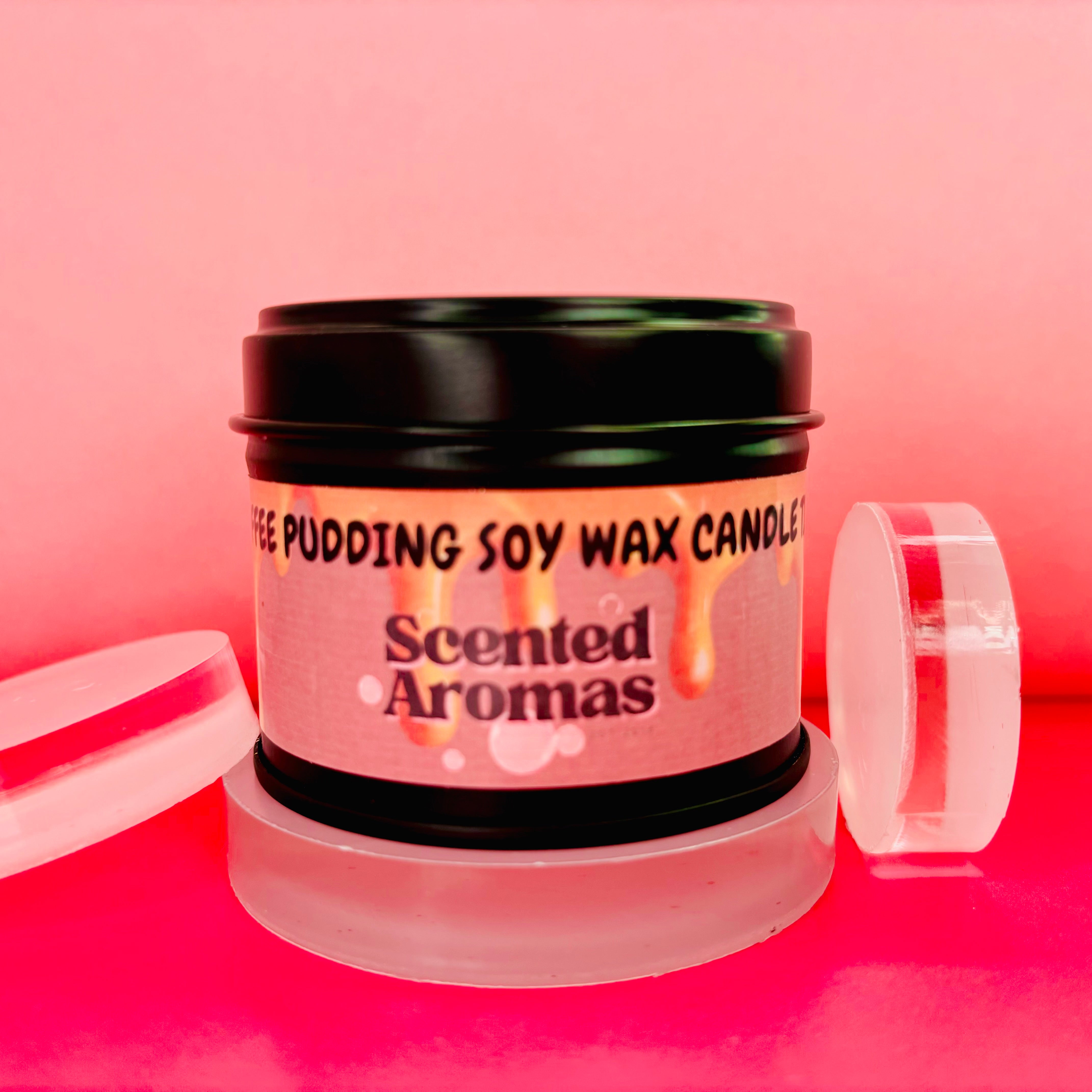 A tin labeled "Sticky Toffee Pudding Candle" from The Soap Gal x rests on a pink surface with clear circular lids beside it, offering long-lasting fragrances for a delightful aroma.