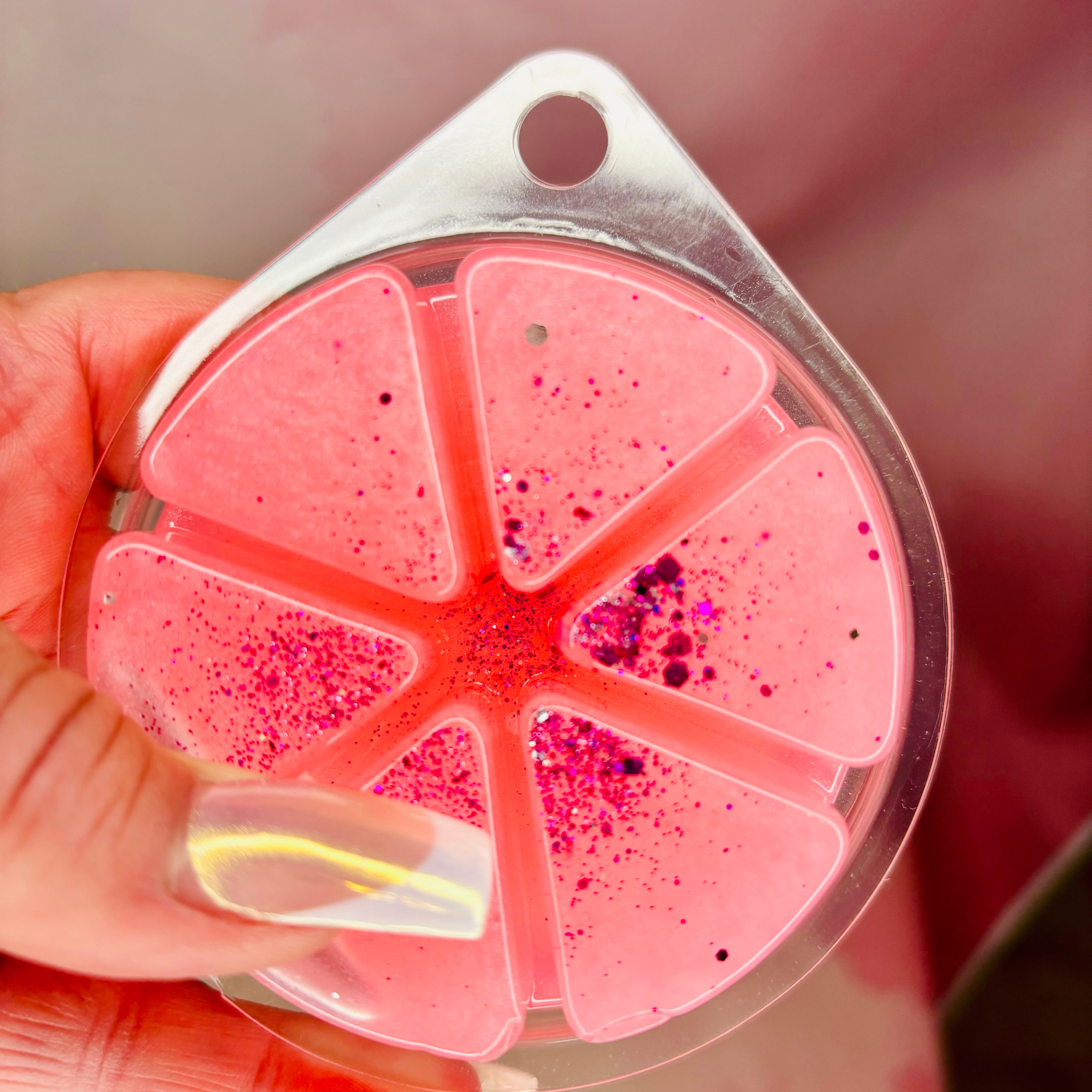 A hand holds a Le Est Belle Gel Wax Melt, crafted by The Soap Gal x, which features a pink glittery design segmented into six triangular sections and encased in a clear plastic mold. Inspired by high-end perfumes, it guarantees a long-lasting fragrance that delights the senses.