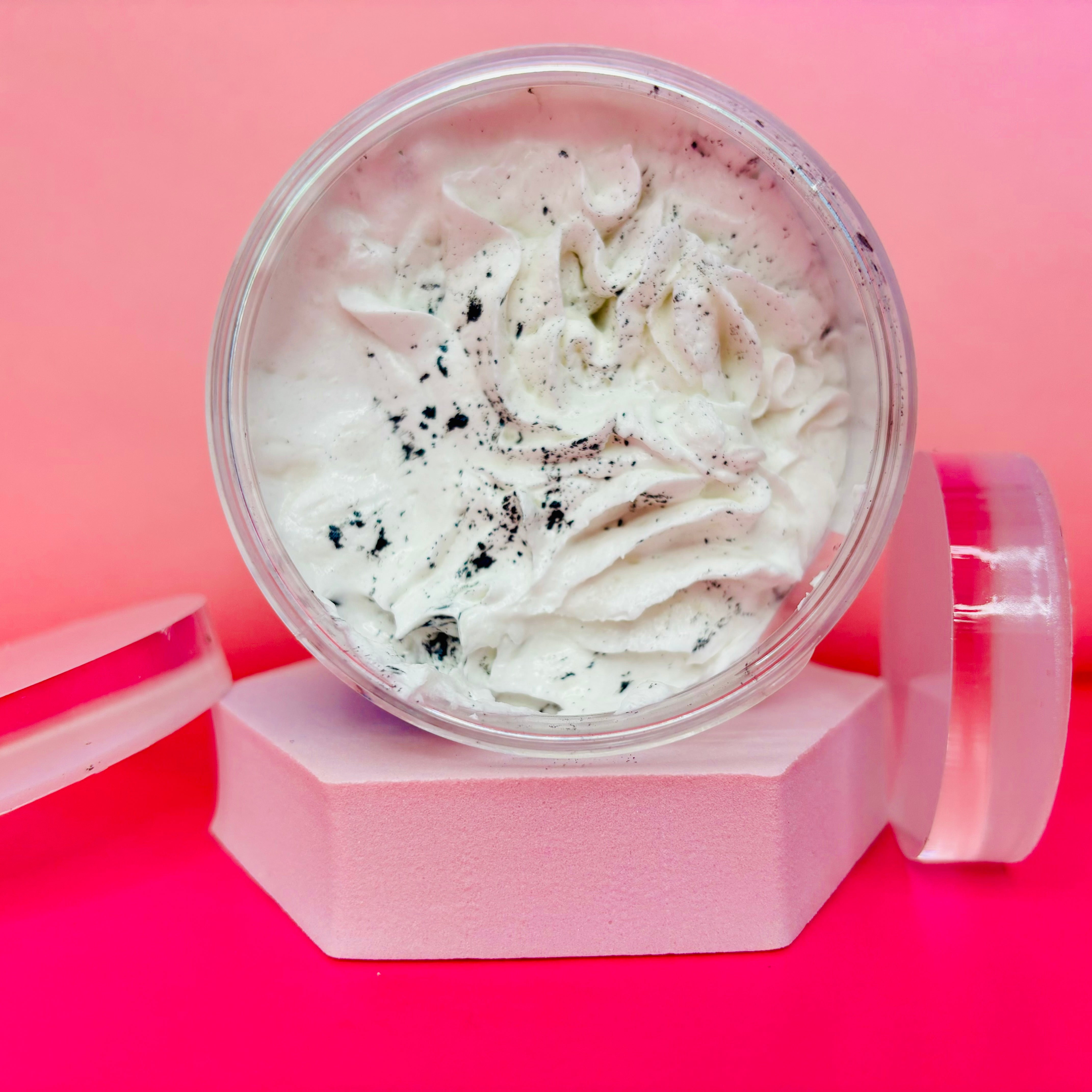 A 200g jar of The Soap Gal x Black Coconut Sugar Body Scrub, boasting a whipped cream texture and exfoliating specks, is displayed with its lid removed, set against a pink and red backdrop to evoke a spa-like ambiance.