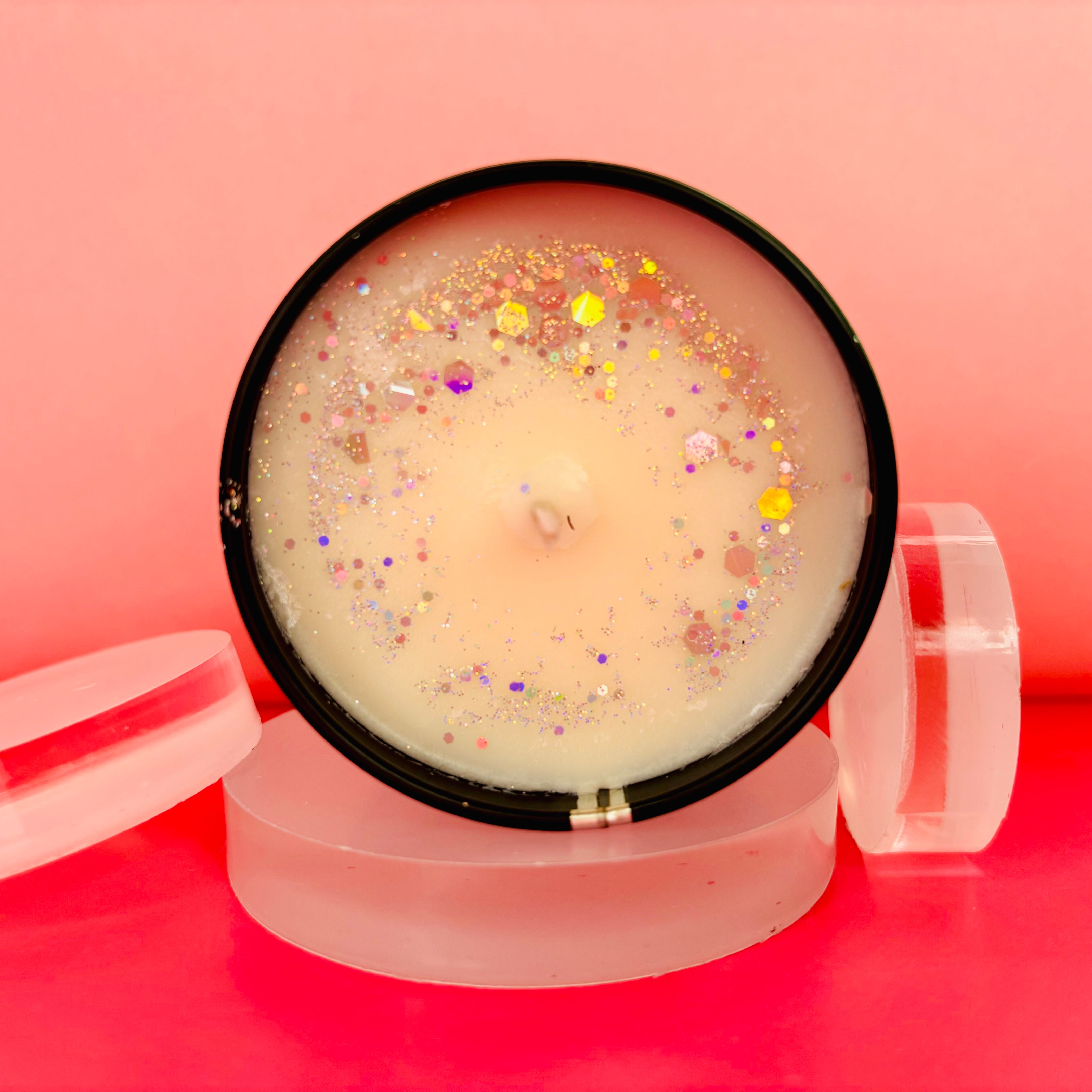 The Soap Gal x's Snow Champagne Candle Tin - 100ml, made from soy wax, is elegantly presented in a black holder adorned with colorful glitter and set on clear circular bases against a pink backdrop.